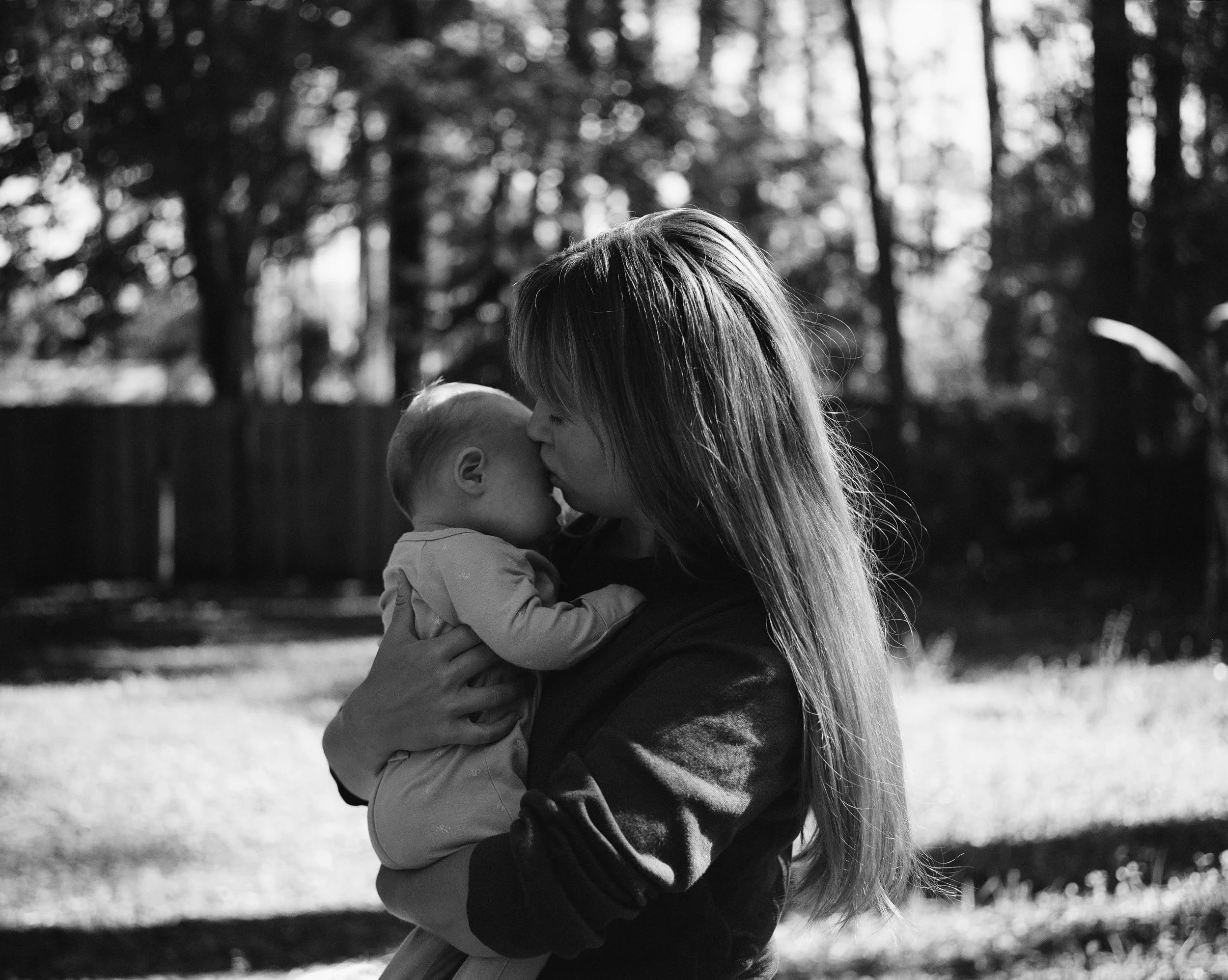 A woman with long hair holding a baby close, kissing the baby's forehead in an outdoor setting with blurred trees in the background.