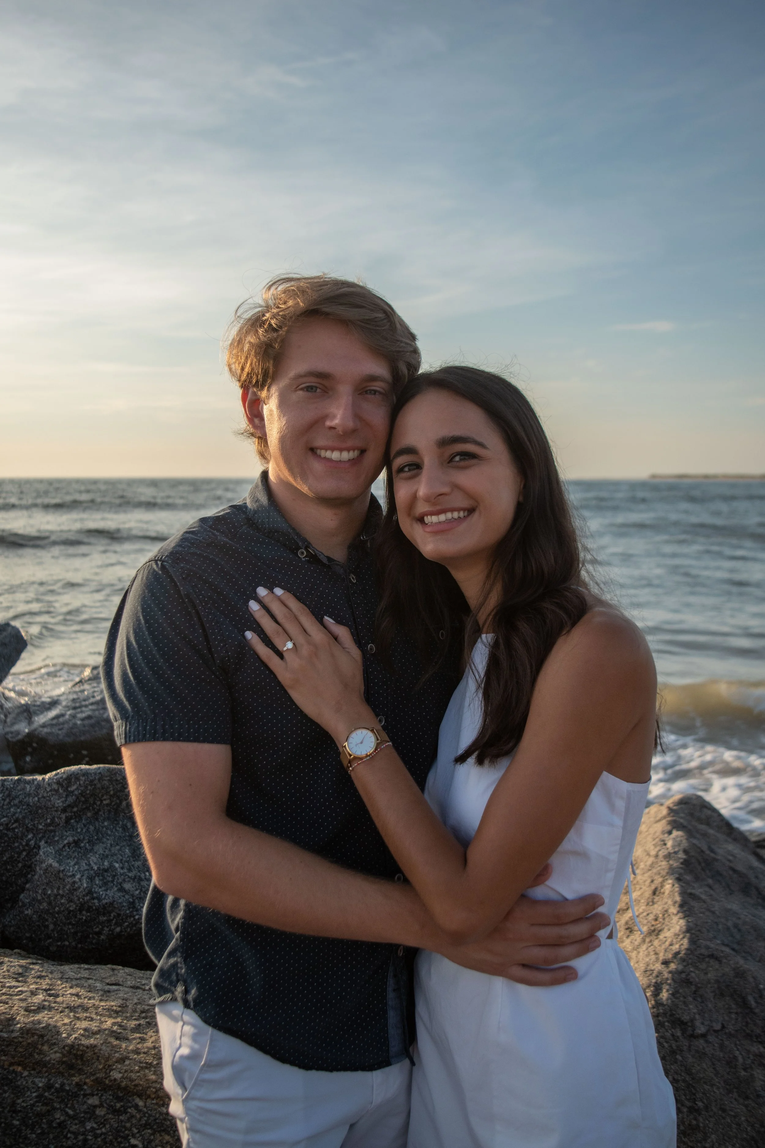 A happy young couple embracing by the ocean on a rocky beach during sunset, smiling at the camera.