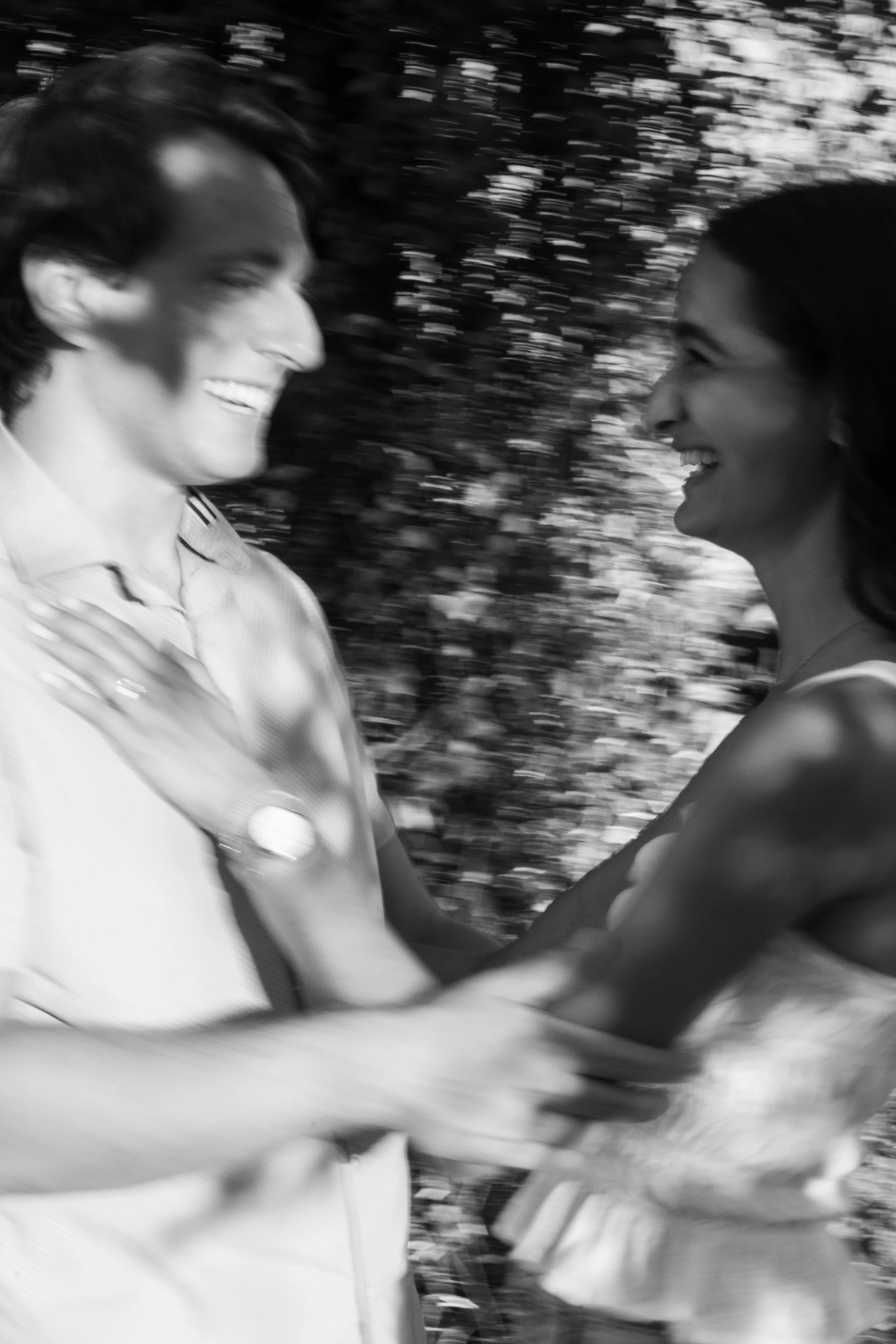 Black and white photo of a man and woman smiling at each other outdoors, with blurred background of trees or bushes, motion-blurred.