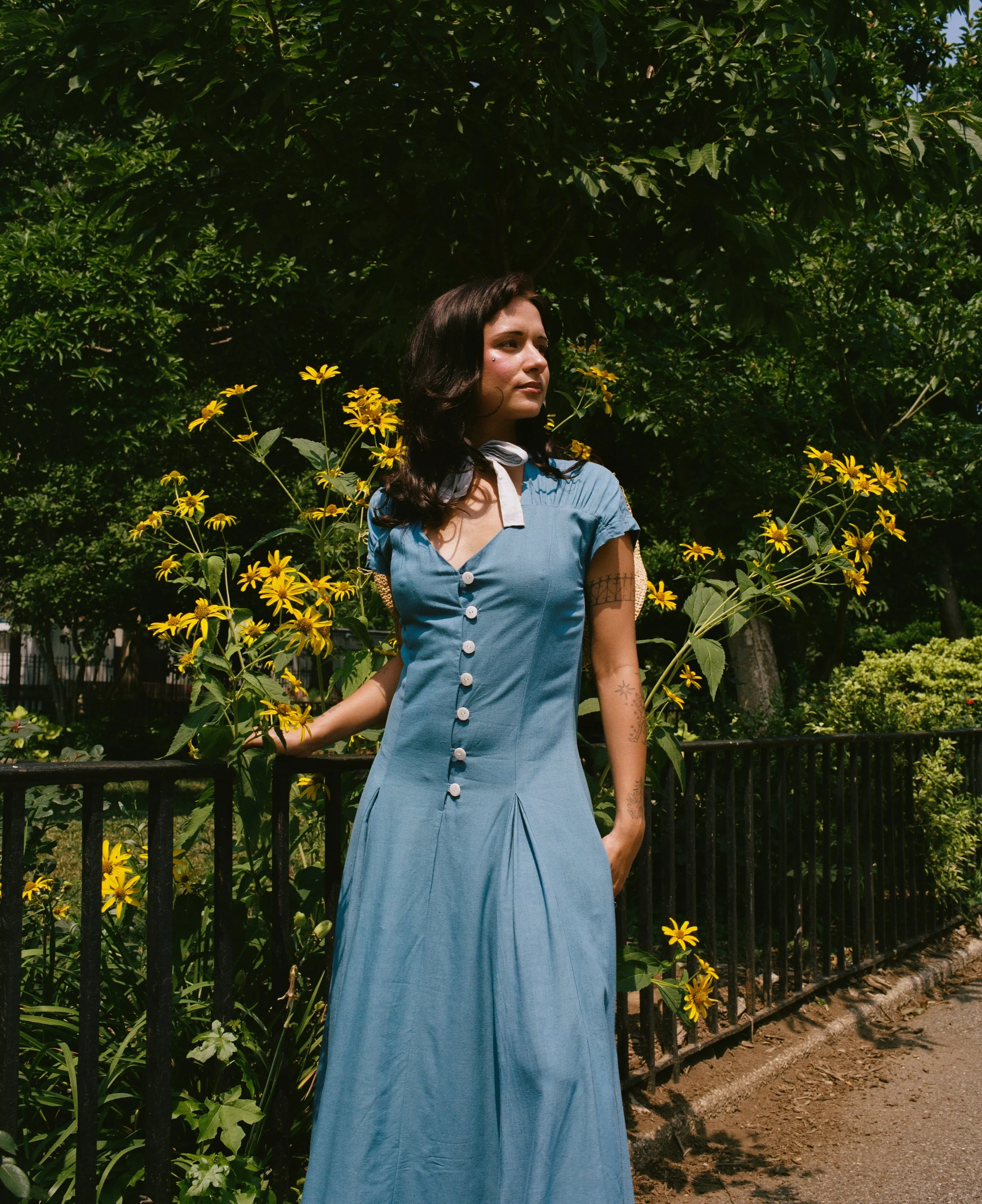 A woman in a blue vintage dress with white buttons, short sleeves, and a white scarf around her neck, standing outdoors in front of lush green trees and yellow flowers, with a black metal fence behind her.
