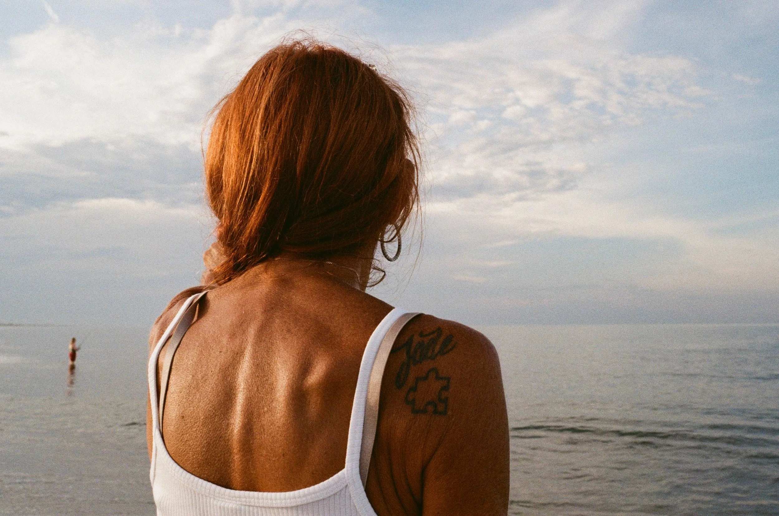 A woman with red hair and tattoos on her arm, wearing a white tank top, looks out at the ocean under a partly cloudy sky.