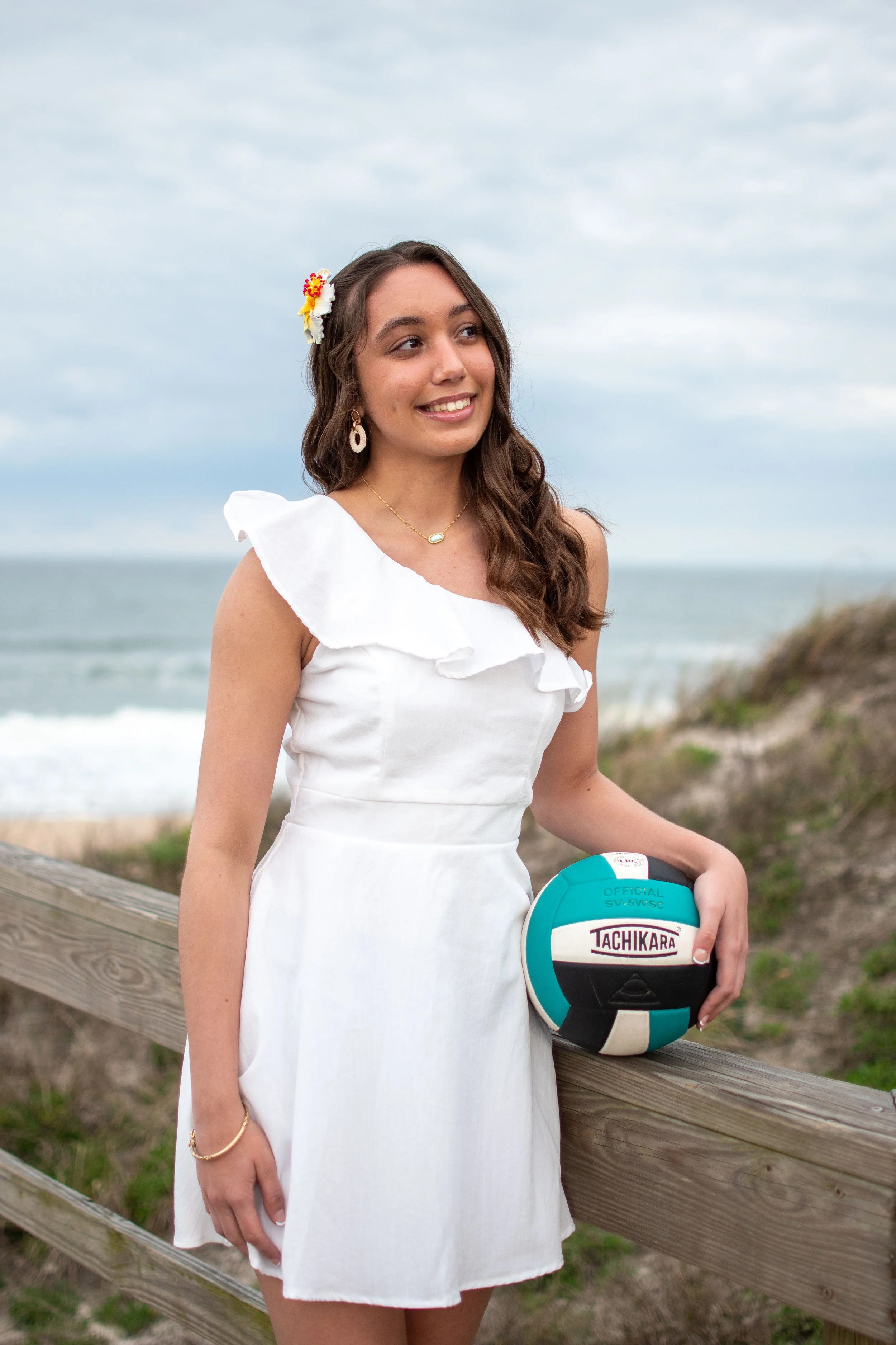 A young woman in a white dress standing near a wooden fence on a beach, holding a turquoise volleyball with white and black details, with the ocean and cloudy sky in the background.