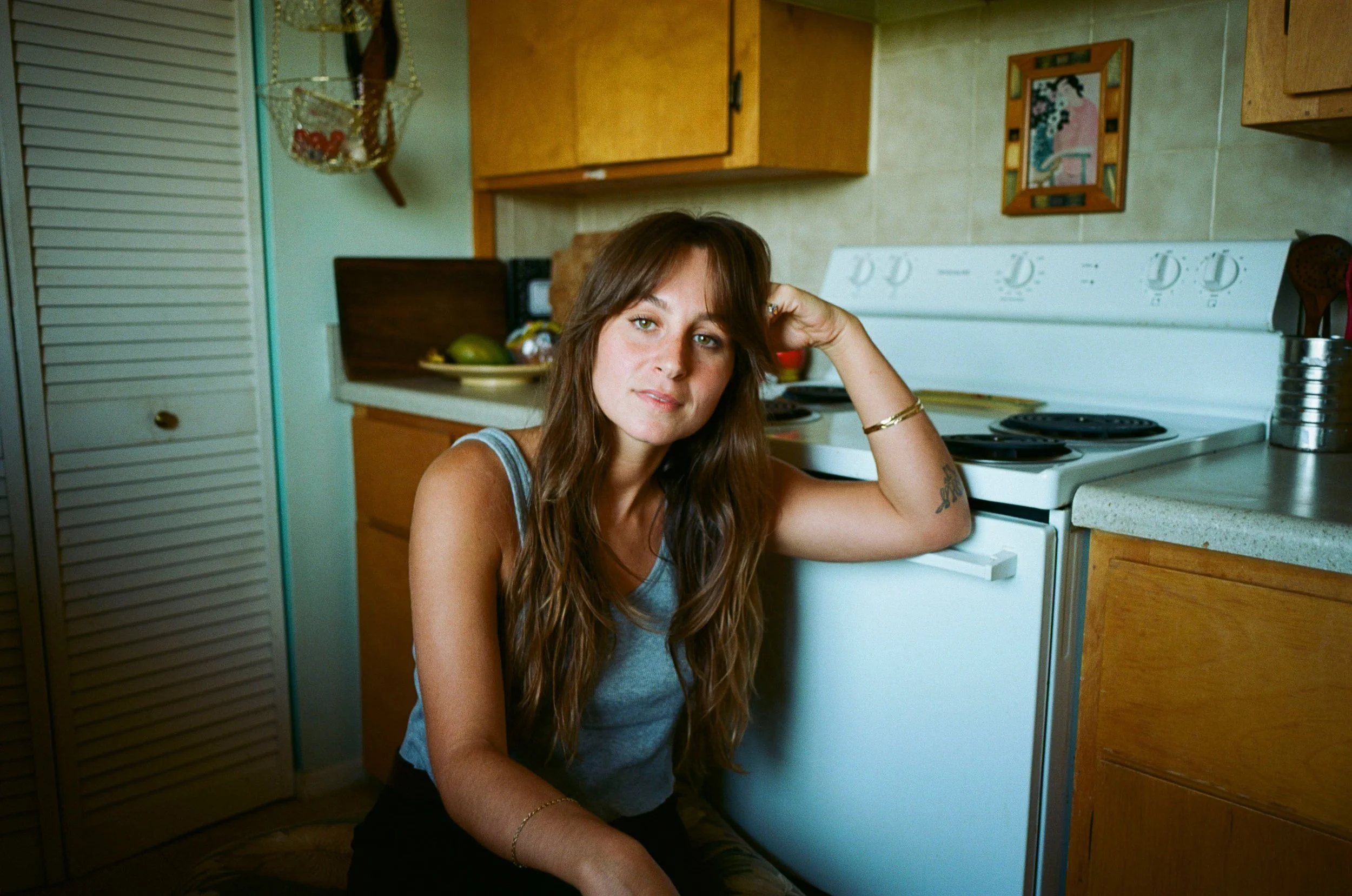 A young woman with long brown hair and a gray tank top sitting on the floor in a kitchen, leaning against a white stove, with one hand resting on her head and looking at the camera.