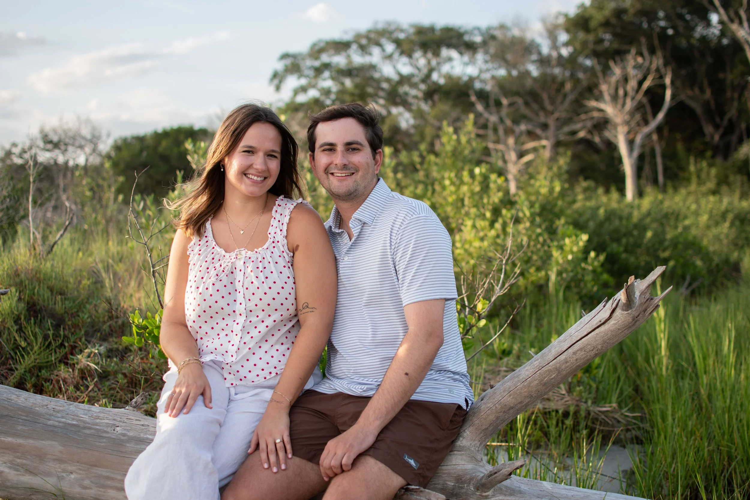 A happy couple sitting on a large fallen tree in a green outdoor setting, smiling at the camera. The woman has shoulder-length brown hair and is wearing a white top with red polka dots, while the man has short dark hair and is wearing a light striped