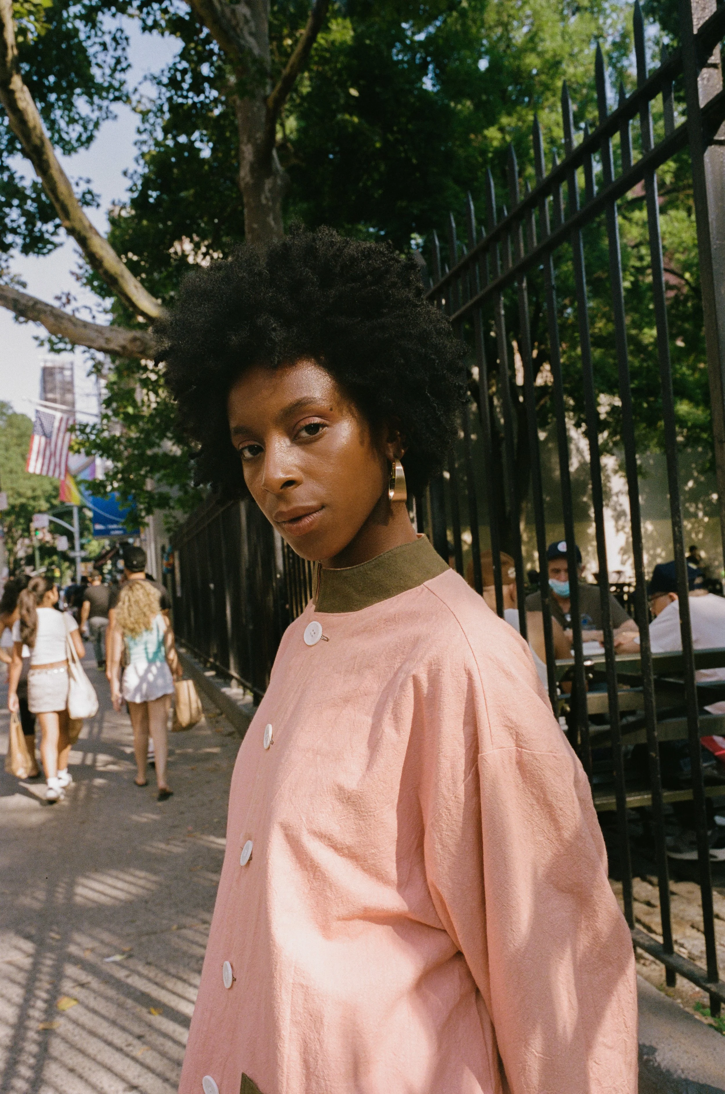 A young Black woman with natural curly hair, wearing a pink coat with white buttons, stands outdoors in an urban setting, with city street, flags, trees, and people in the background.