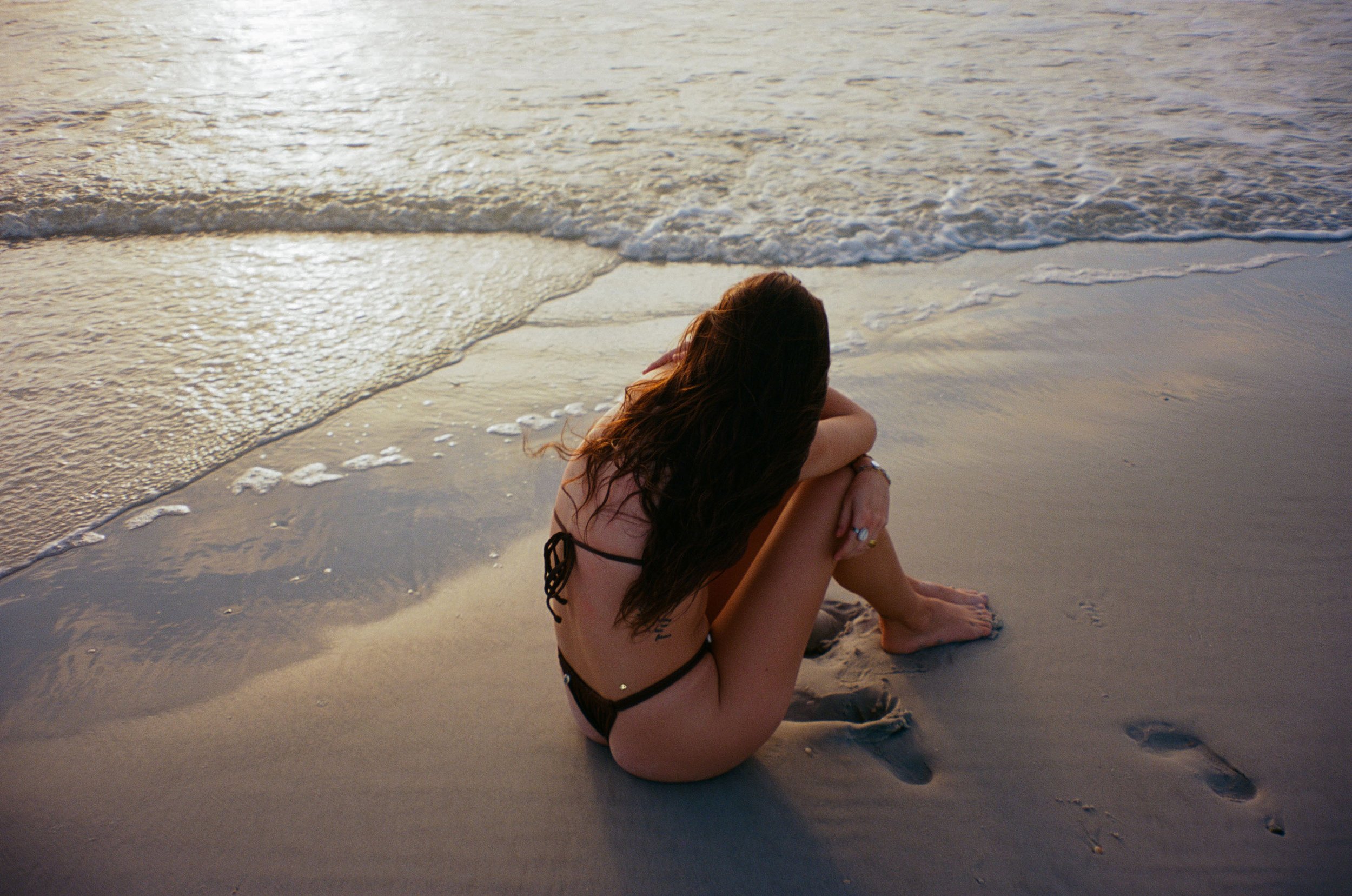 A woman with long hair sitting on the beach at sunset, facing the ocean, with her arms around her knees.