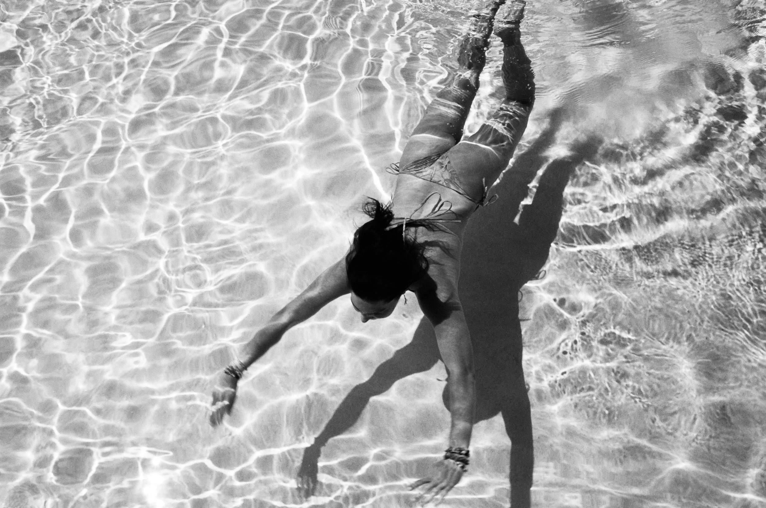 A person swimming underwater in a pool, captured in black and white, with ripples and reflections on the water surface.