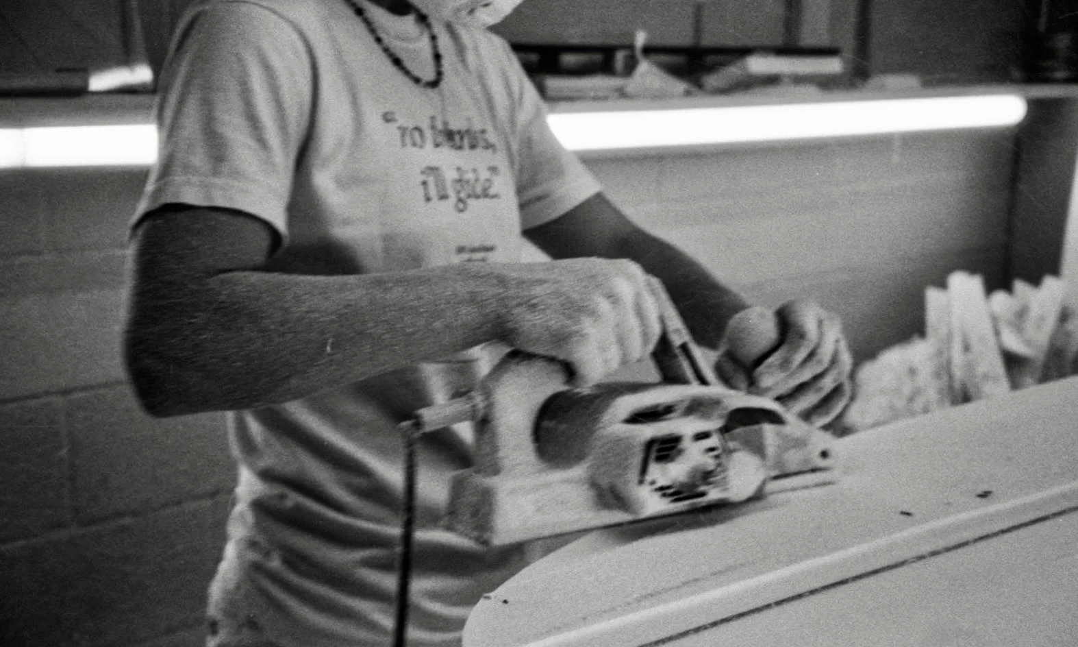 A person operating a handheld electric saw while working in a workshop or hardware store. The person is wearing a T-shirt with text on it and has a beaded necklace. The background includes shelves with various items.