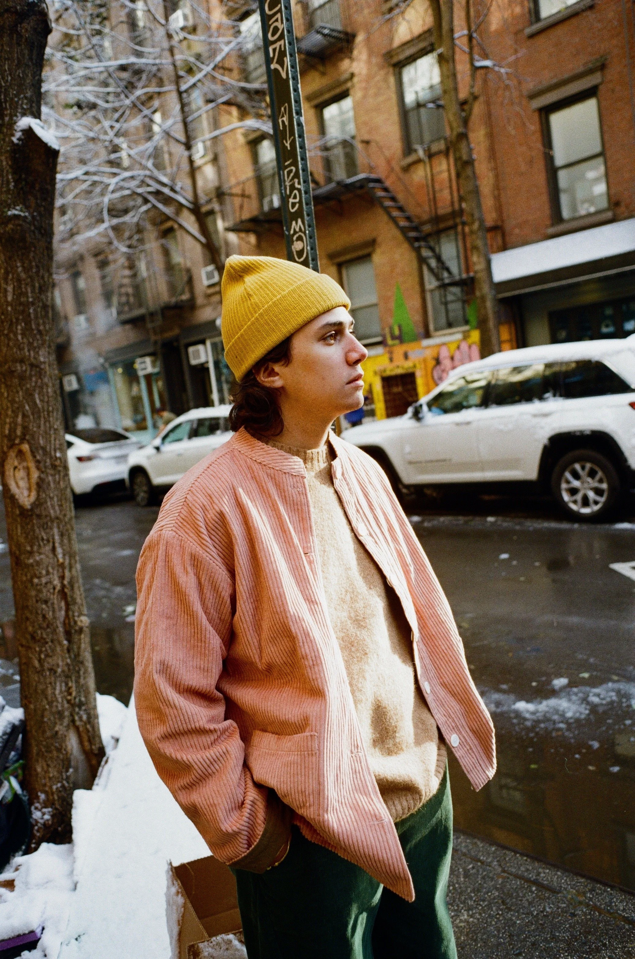 A person with shoulder-length dark hair wearing a yellow beanie, pink jacket, and beige sweater standing on a snowy city street with parked cars and brick buildings in the background.
