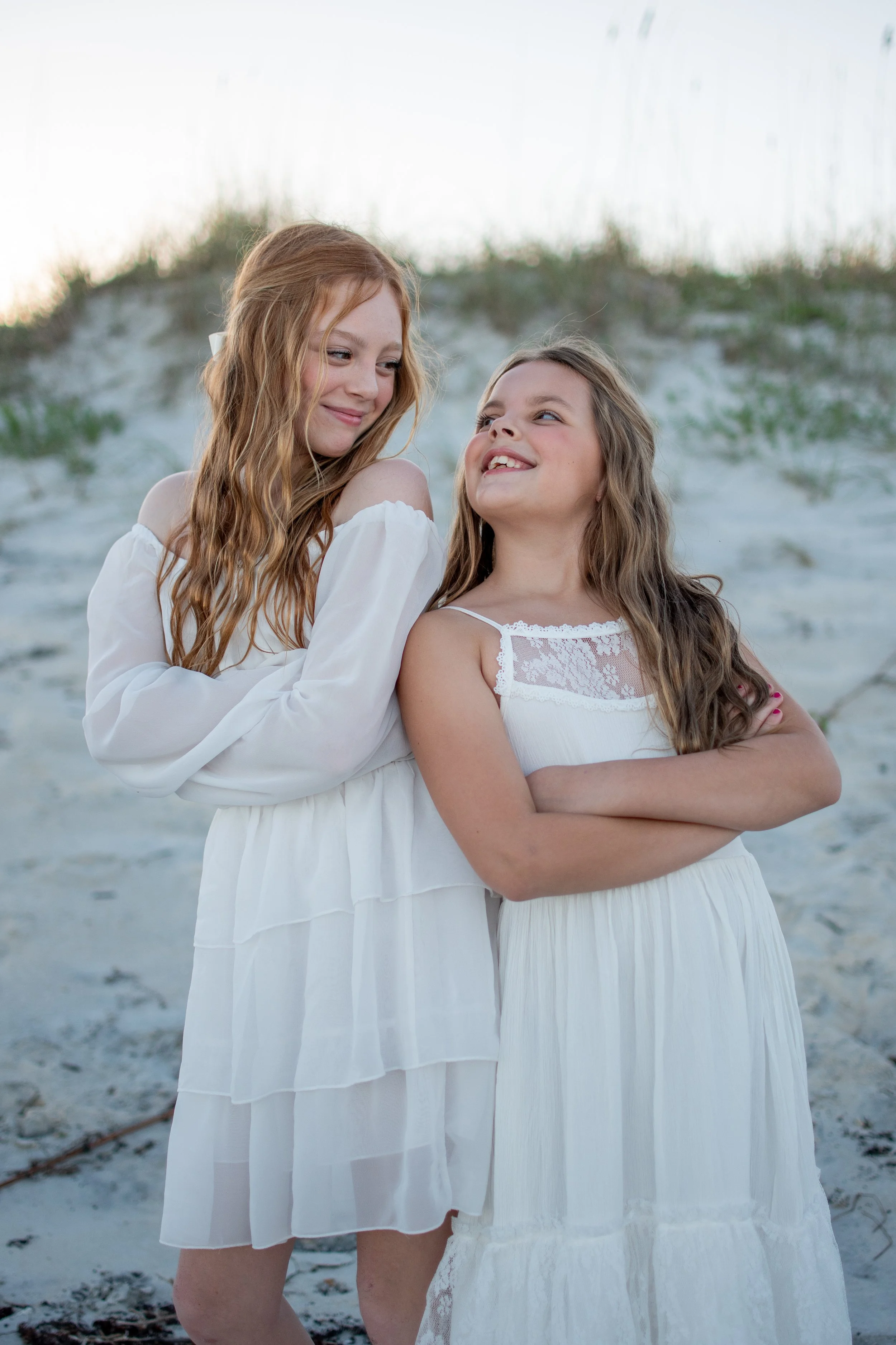 Two young girls wearing white dresses stand close together on a beach with sand and sparse grass in the background, smiling and looking at each other.