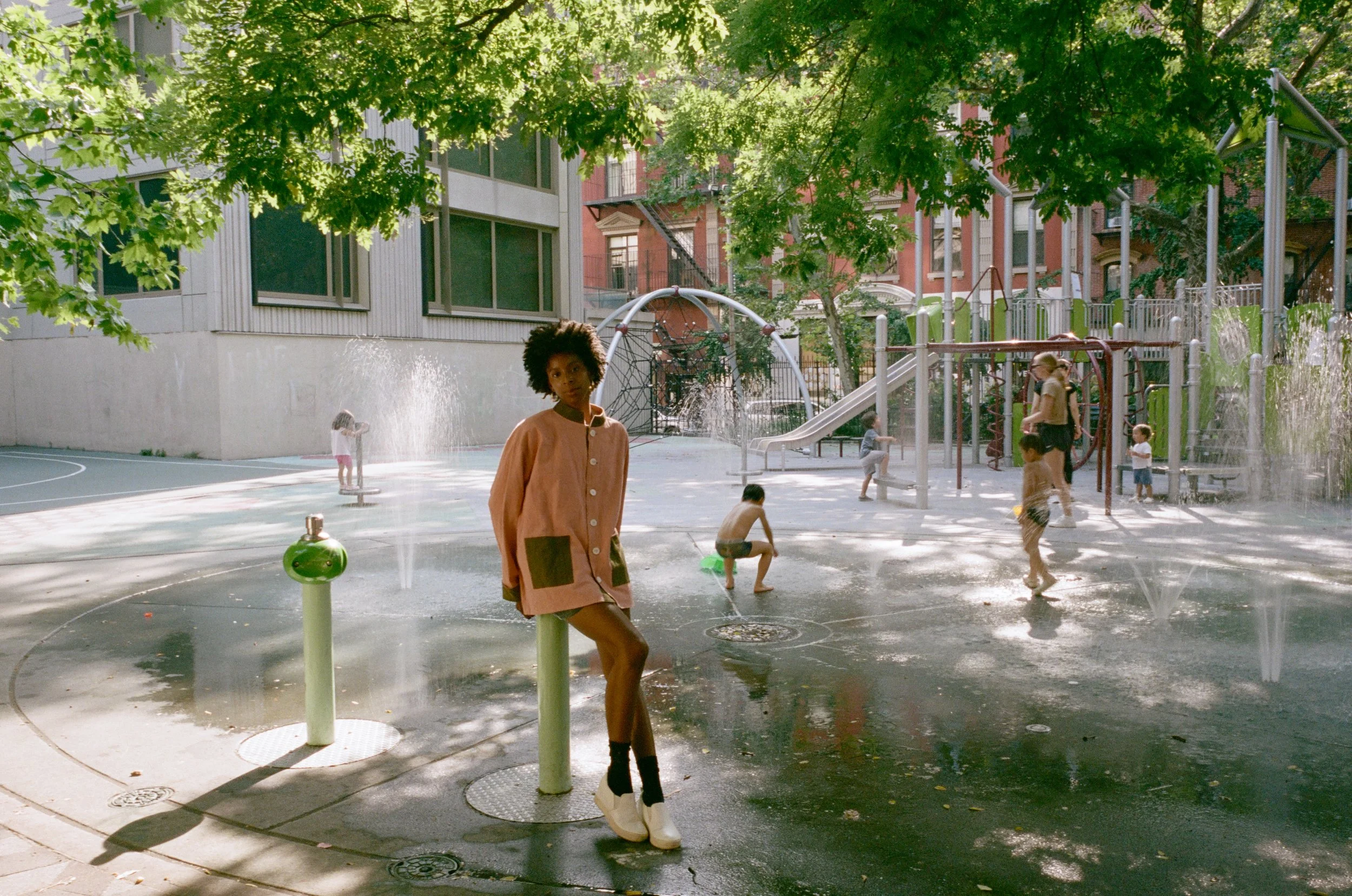 Children playing in a splash pad area with water sprays at a city park, surrounded by green trees and residential buildings.