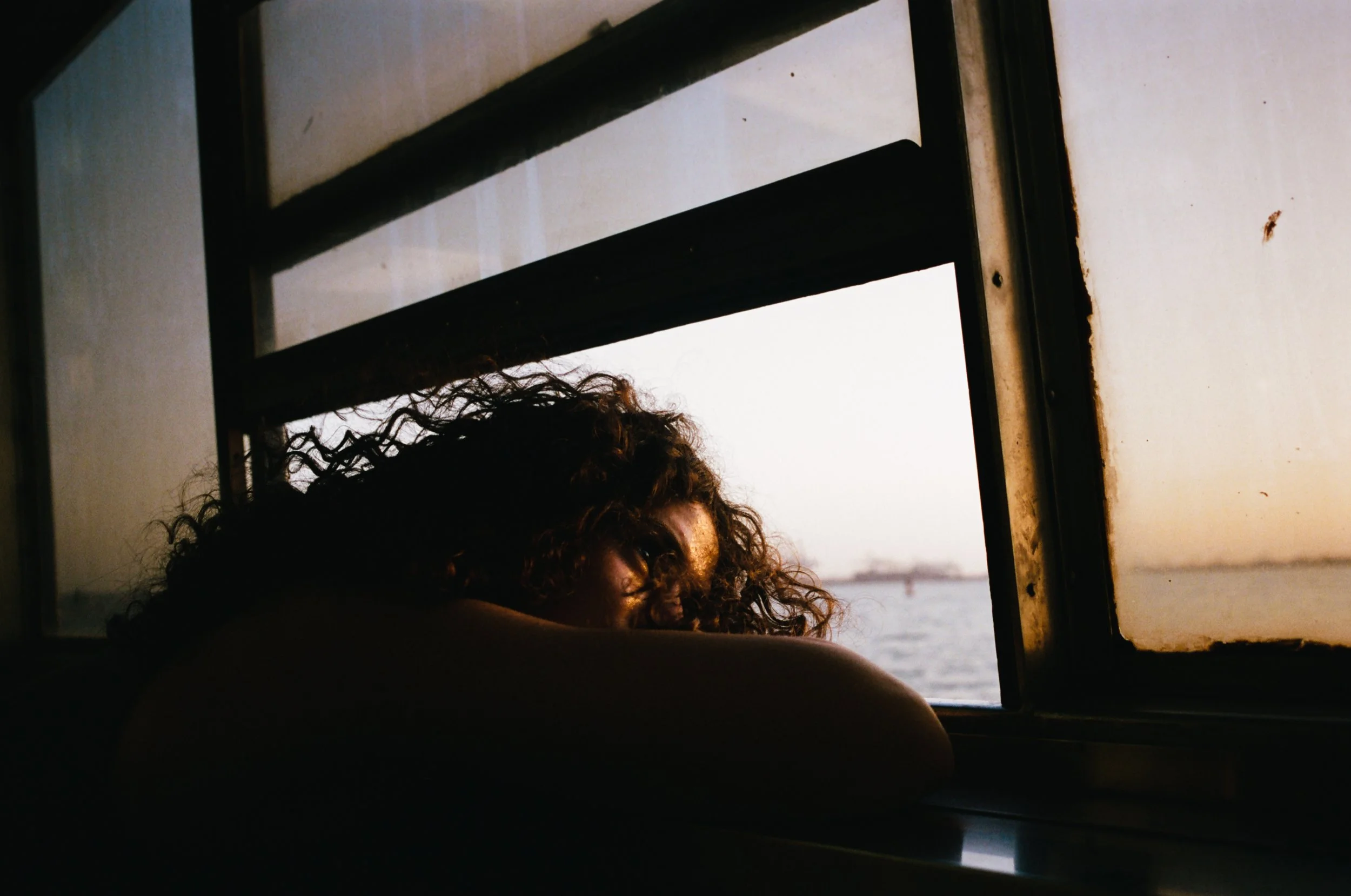 A person with curly hair leaning on a surface, looking out of a window during sunset or sunrise over the water with a bridge in the background.