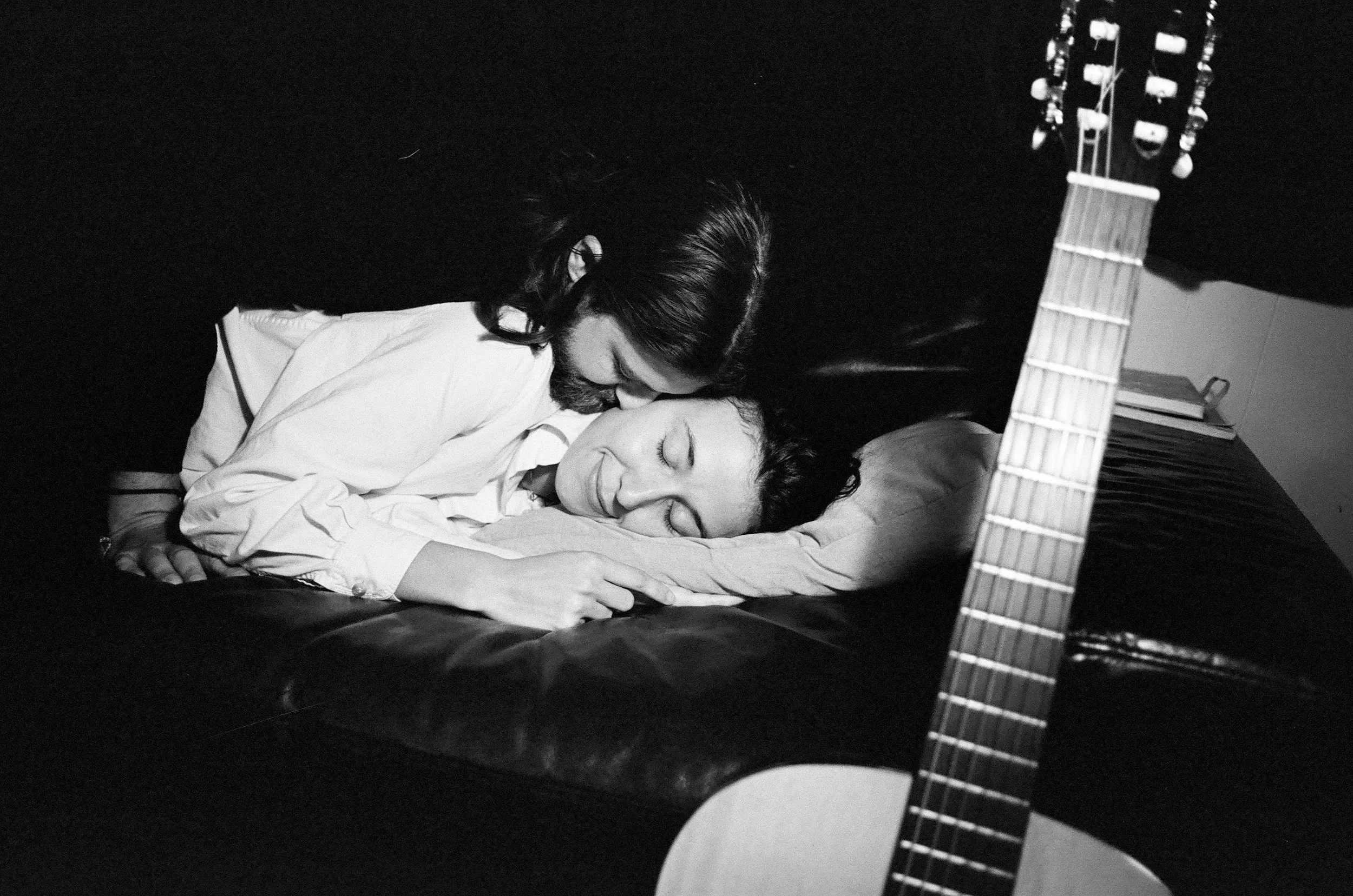 A black and white photo of a couple cuddling and sleeping together. The woman is resting her head on the man, and they both appear peaceful and content. A guitar is partially visible in the foreground.