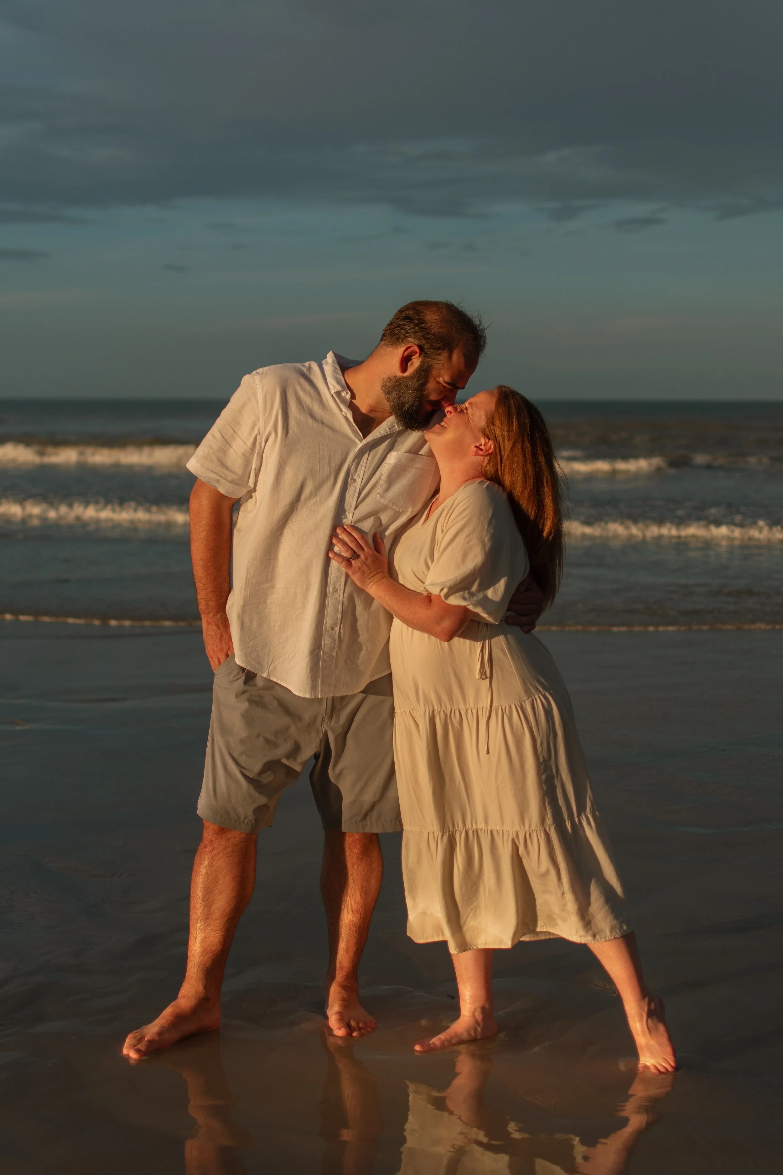 A couple standing at the beach, close together, with the ocean and clouds in the background, during sunset.