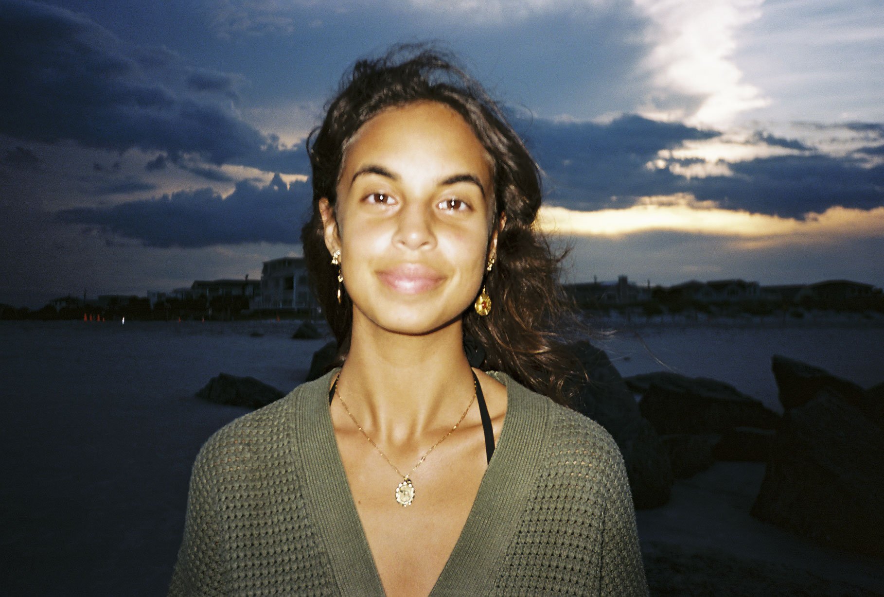 A woman with dark hair smiling outdoors at dusk, standing on a beach with rocks and buildings in the background, dark clouds and a sunset sky.