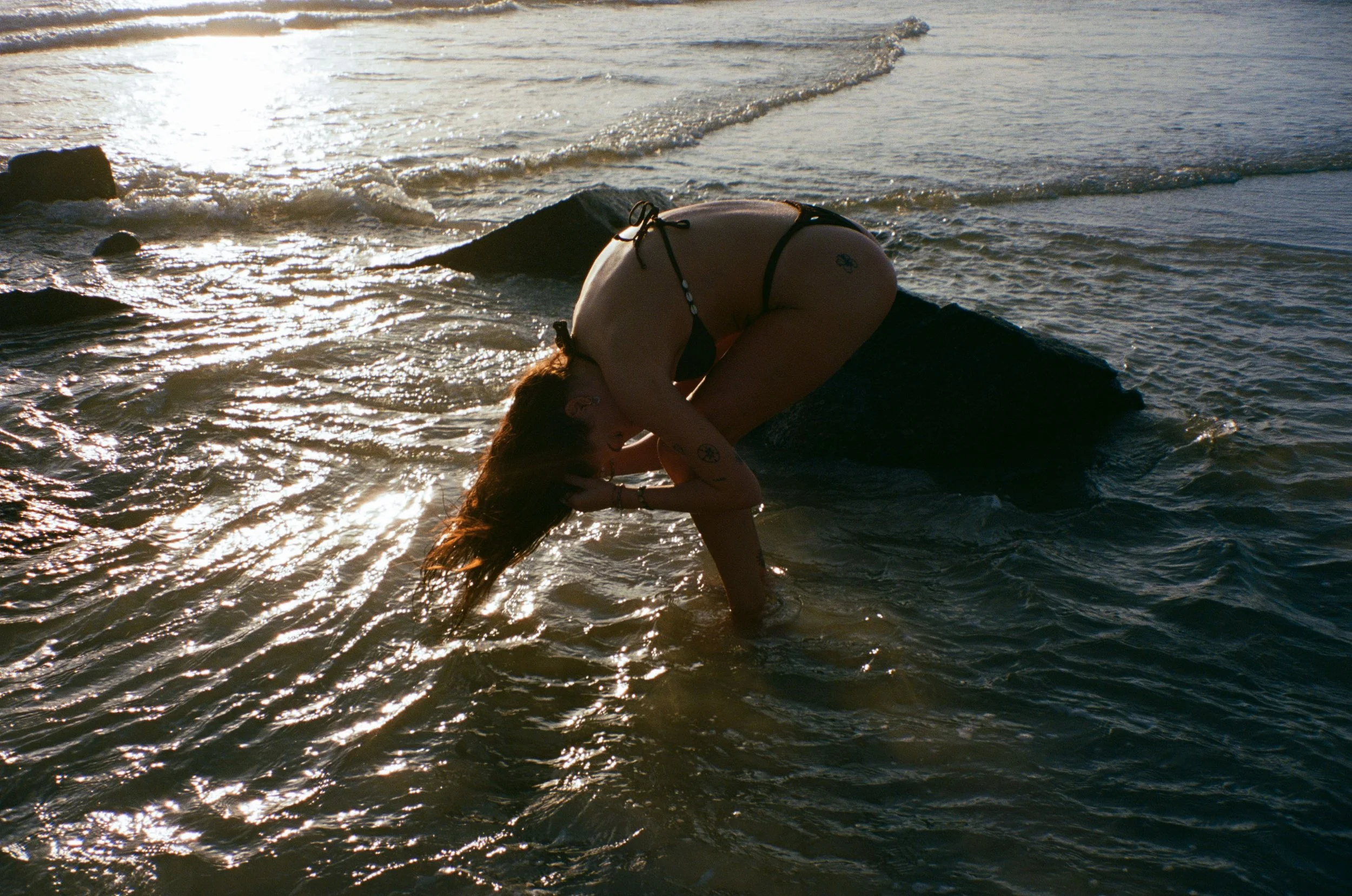 A person with long hair wearing a bikini, bending forward on a large rock at the edge of the water, with waves and sunlight in the background.