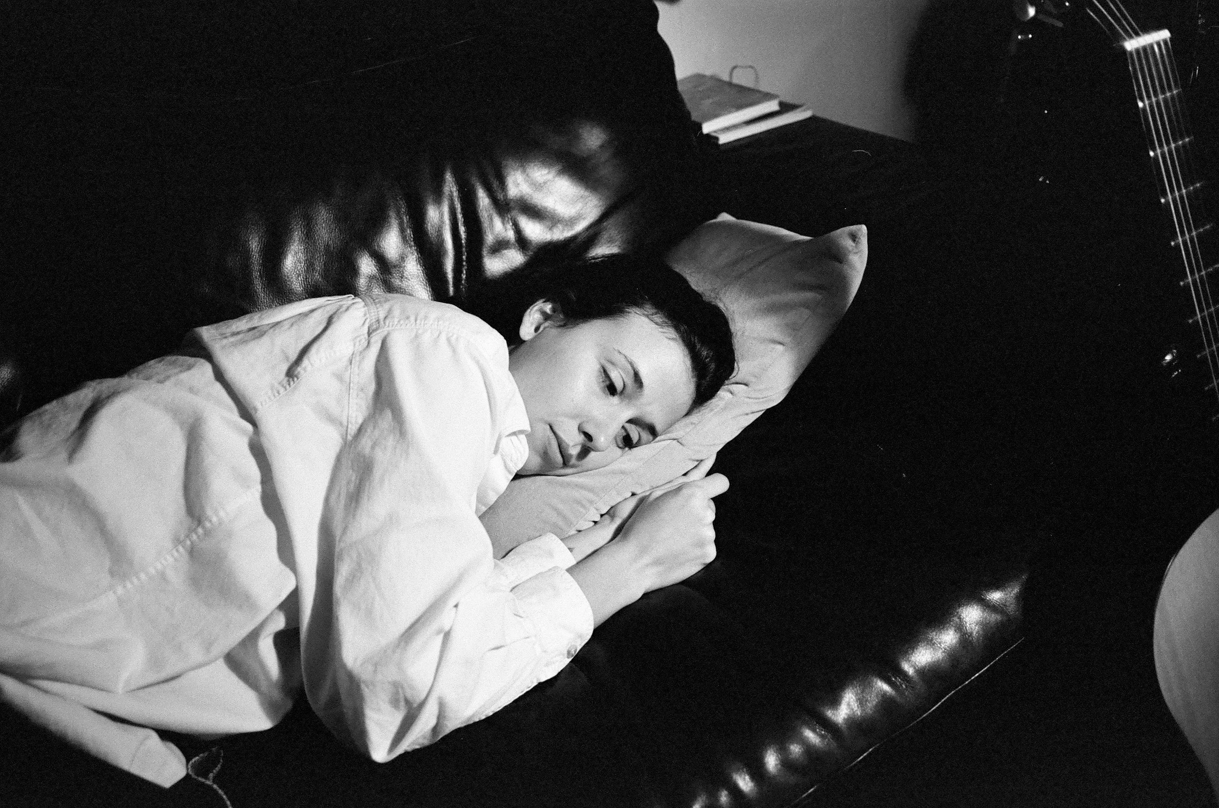 A woman lying on a piano bench resting her arm on the piano, with her eyes closed, wearing a light-colored shirt and a man with dark hair and a leather jacket leaning over her, in a black-and-white photo.