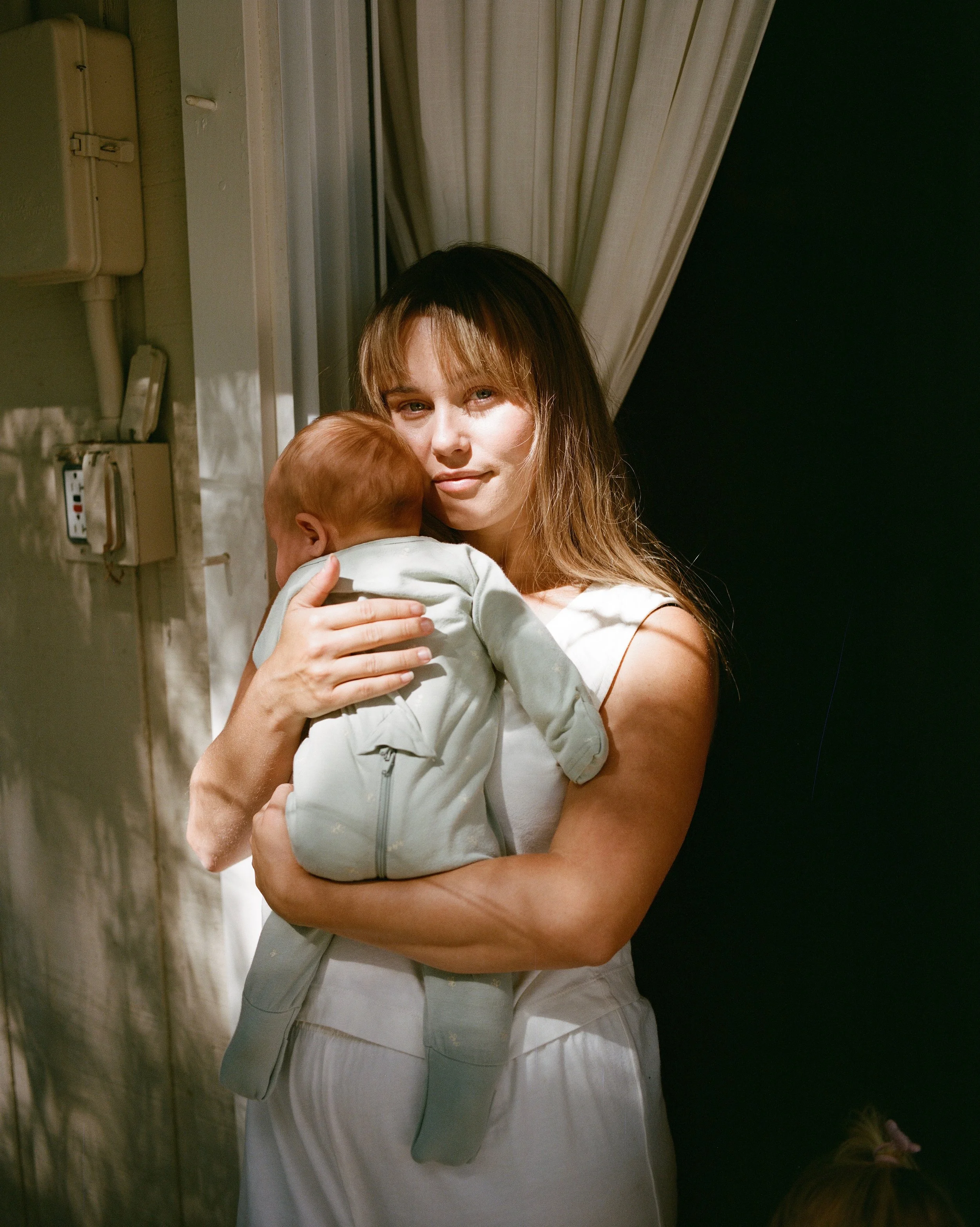 A woman holding a baby indoors, near a curtain and electrical panel, with a dark background to the right.