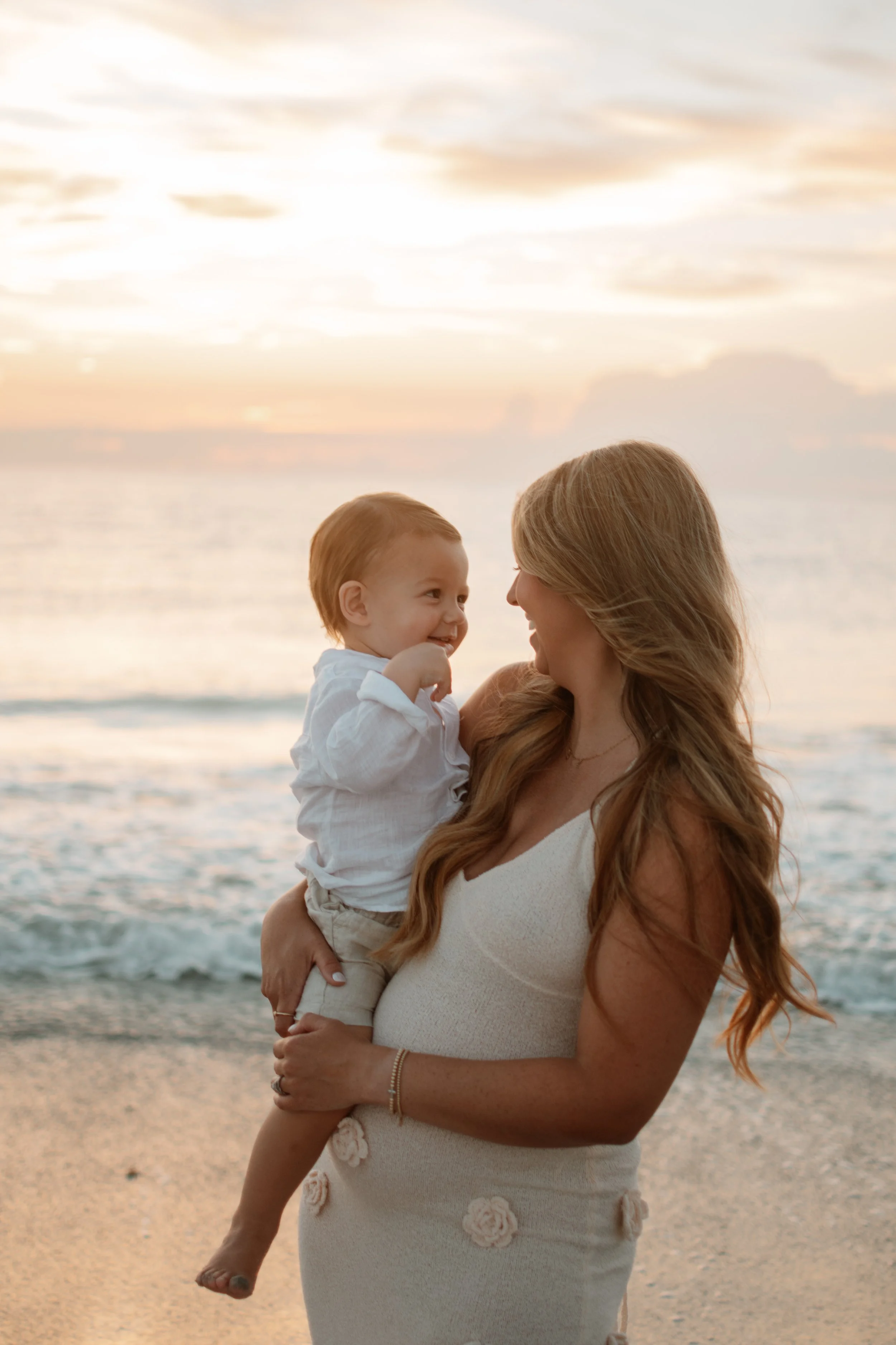 A woman with long, wavy hair holding a young boy on a beach at sunset, both smiling and looking at each other, with the ocean and a cloudy sky in the background.