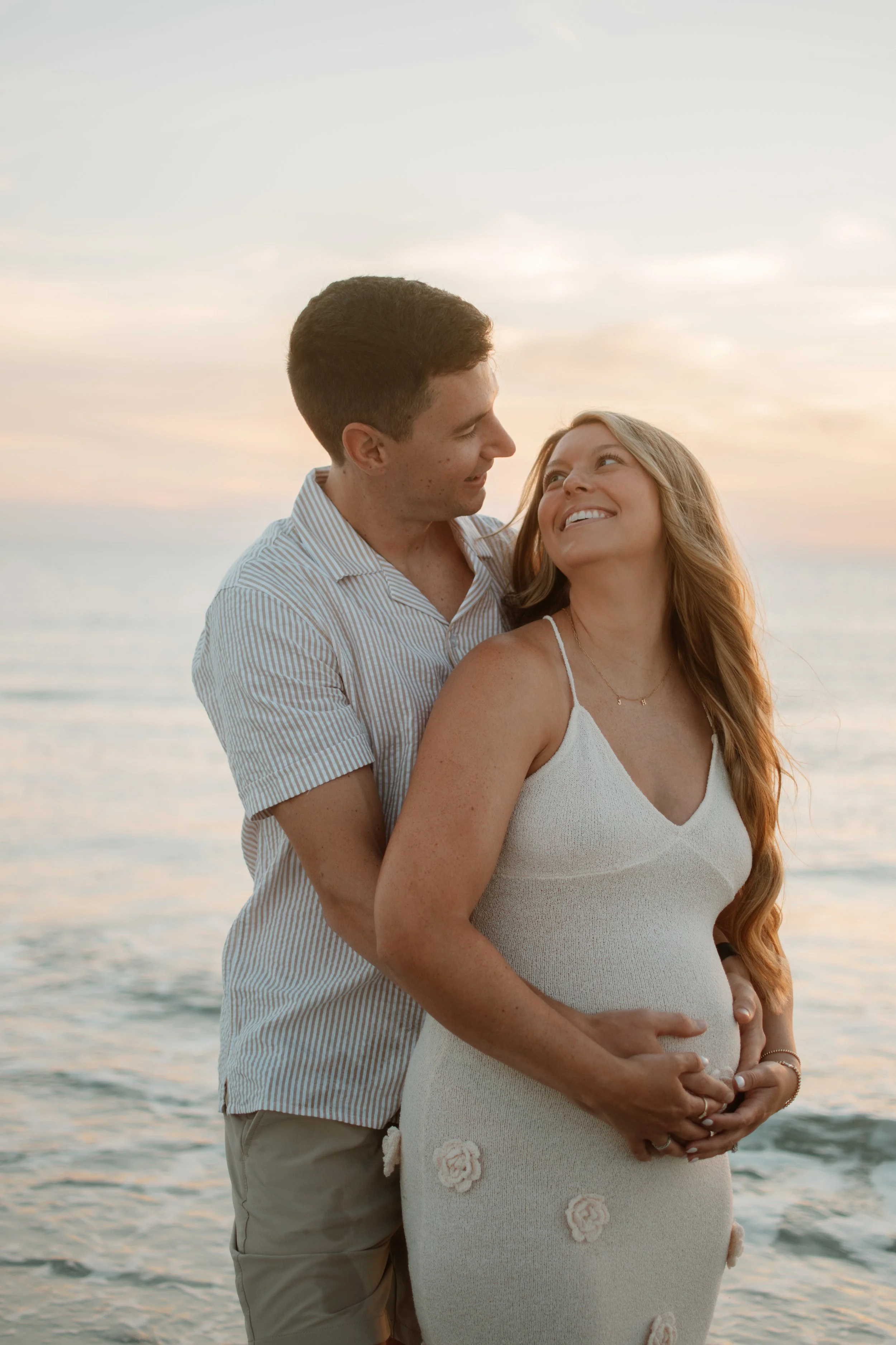 A pregnant woman and her partner at the beach during sunset, both smiling and lovingly looking at each other.