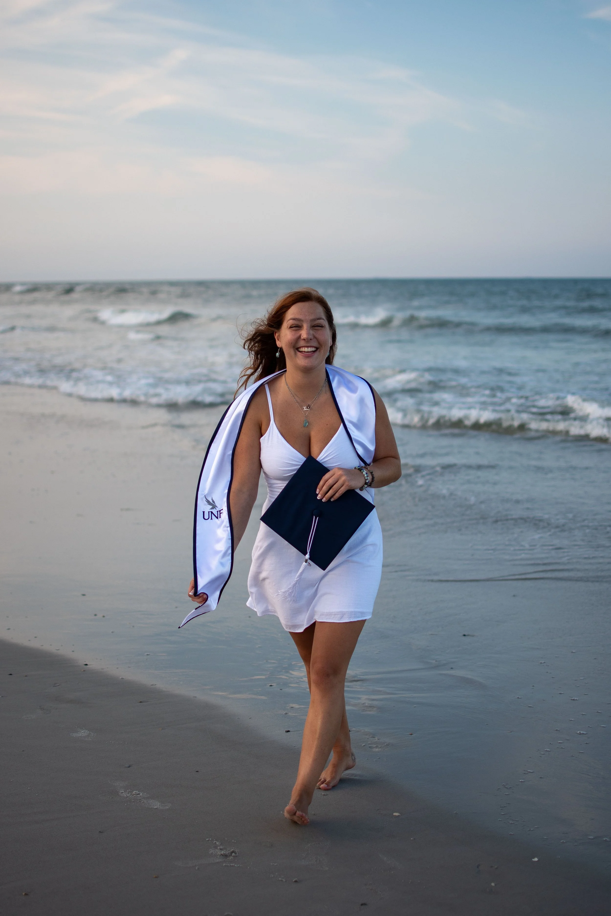 A young woman in a white dress walking barefoot on the beach holding a graduation stole and diploma, with the ocean and cloudy sky in the background.