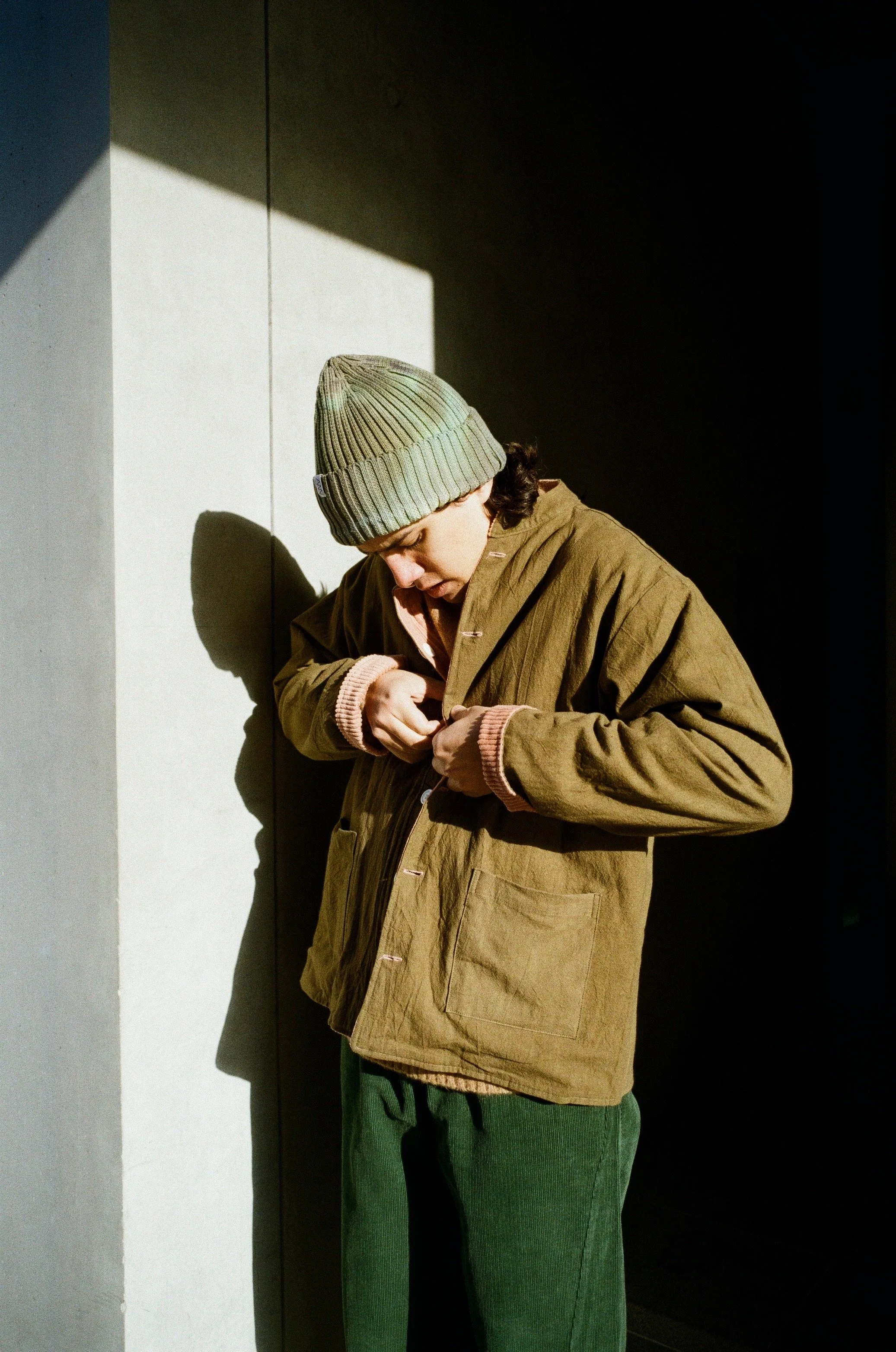 A person wearing a gray beanie, a brown coat, and gray pants standing against a wall with contrasting shadows and light. The person is looking down, adjusting something with their hands.