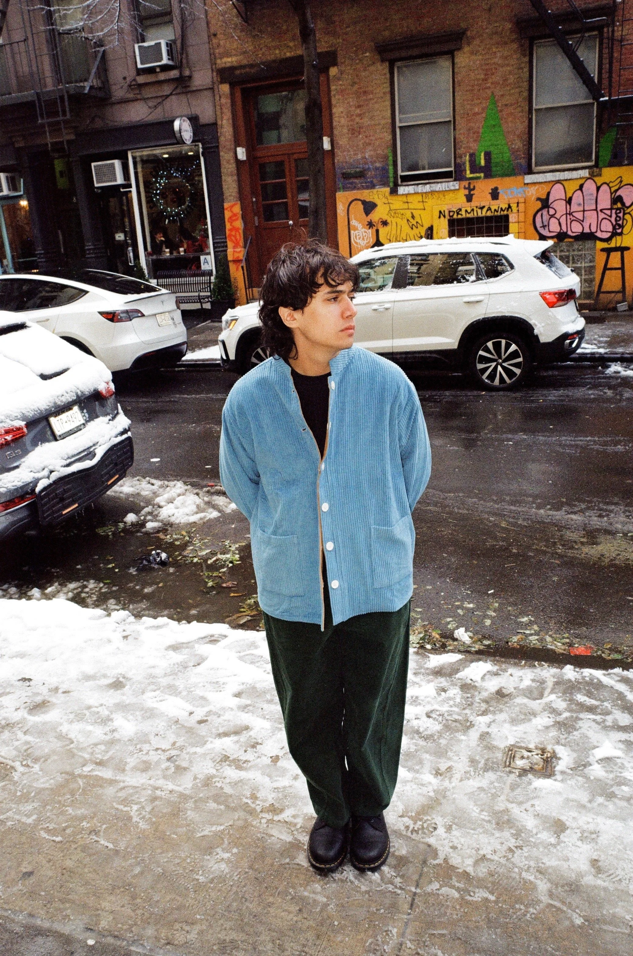 A young man in a blue jacket and black pants standing on a snow-covered street in an urban neighborhood with graffiti-covered walls, parked cars, and storefronts in the background.