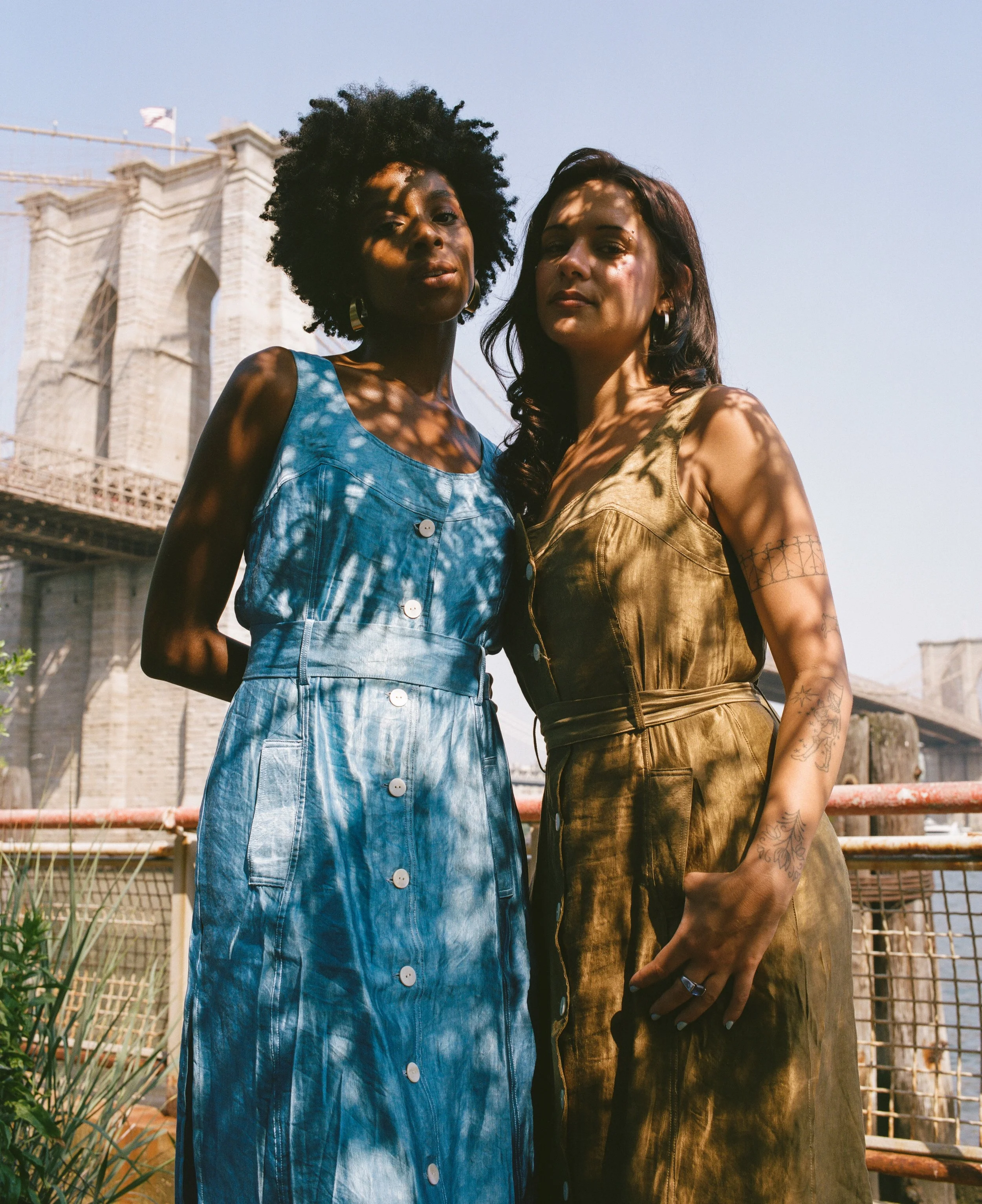 Two women standing close together outdoors in front of the Brooklyn Bridge, casting shadows on their faces from nearby trees, during daytime.
