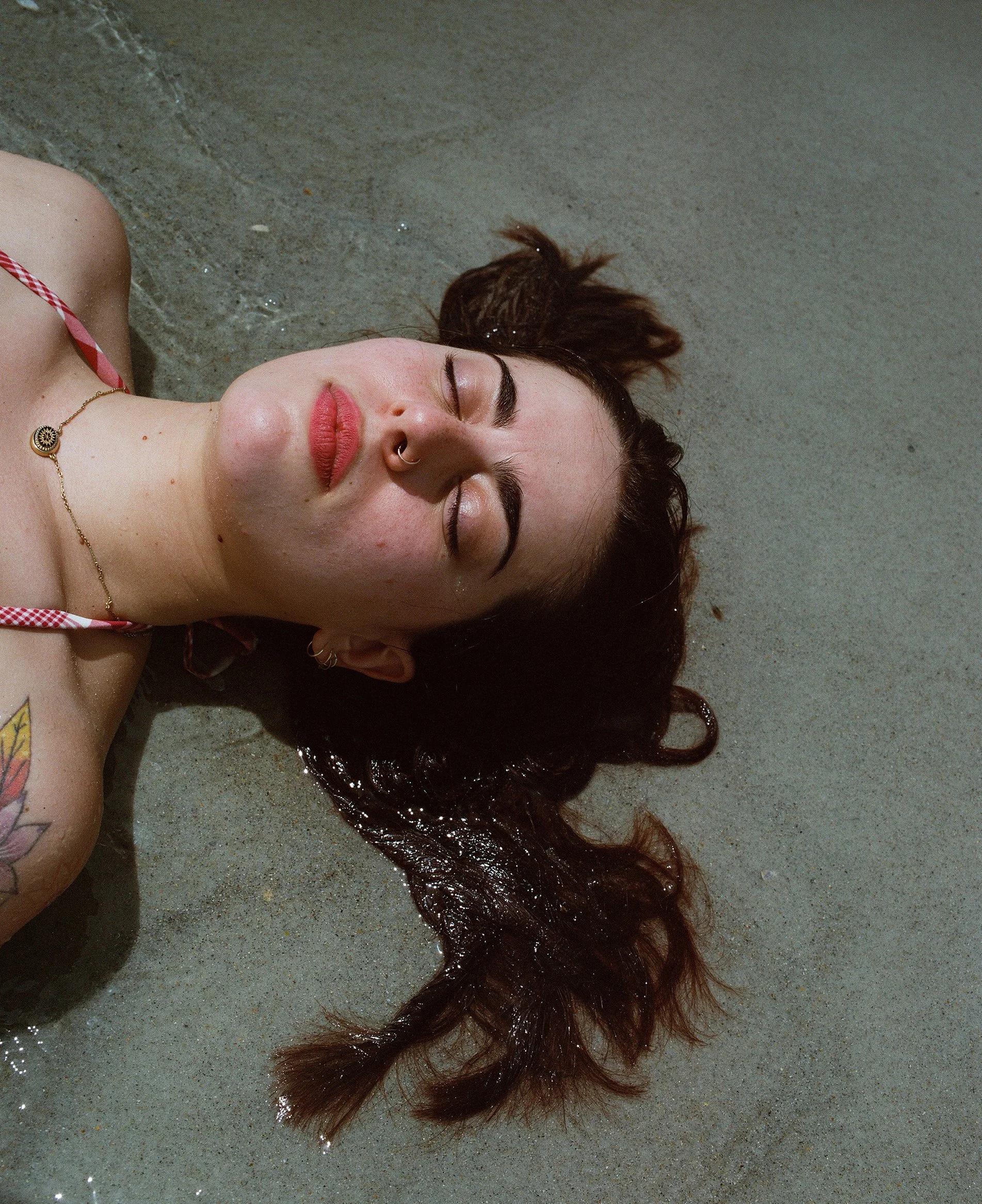 A woman with brown hair and a nose ring lying on a sandy beach with eyes closed, wearing a pink and white checkered swimsuit and gold jewelry.