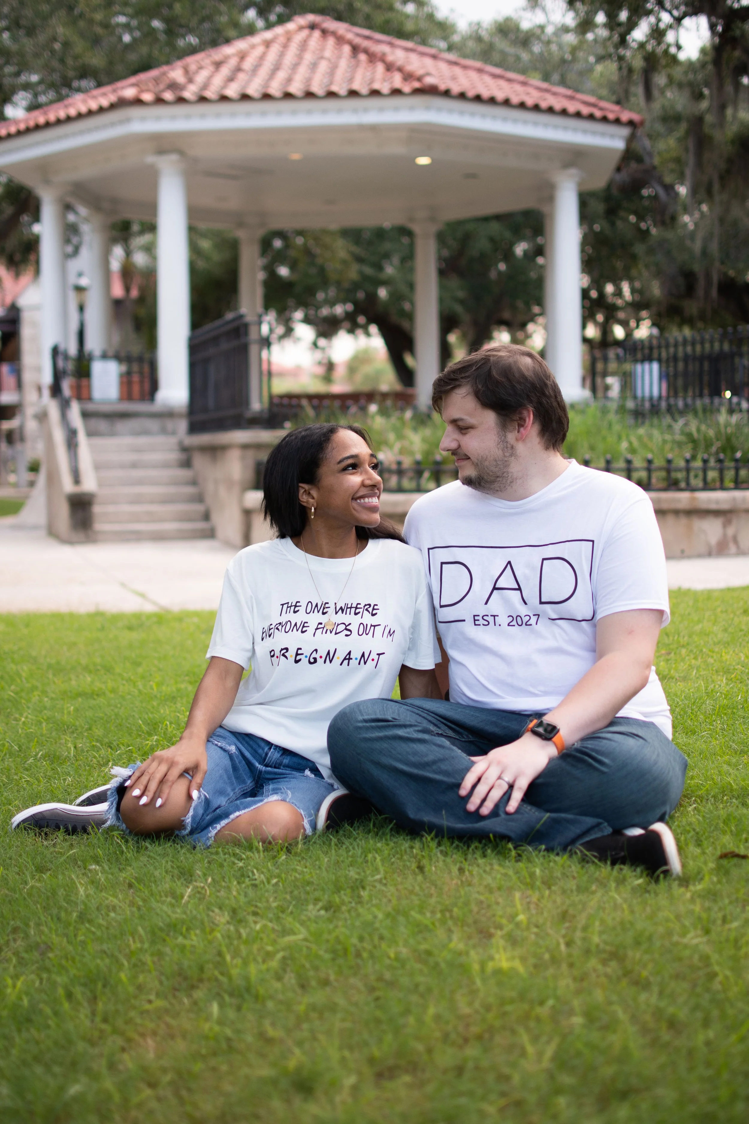 A couple sitting on grass in front of a gazebo, smiling at each other. The woman is wearing a white T-shirt with printed text, and the man is wearing a white T-shirt with text that says 'DAD' and 'Established 2027'.