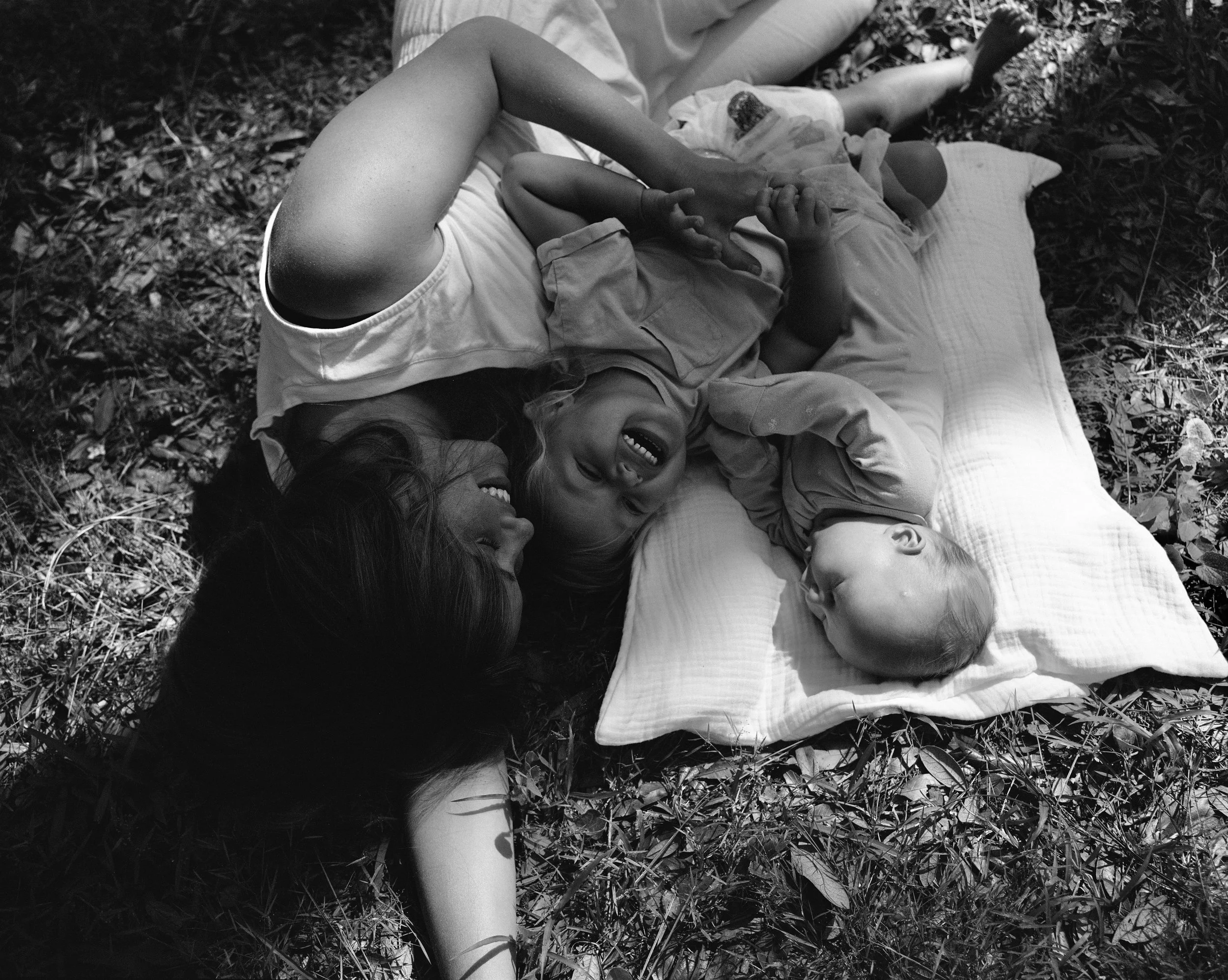 A girl lies on a picnic blanket with children, smiling and laughing, outside in a wooded area.