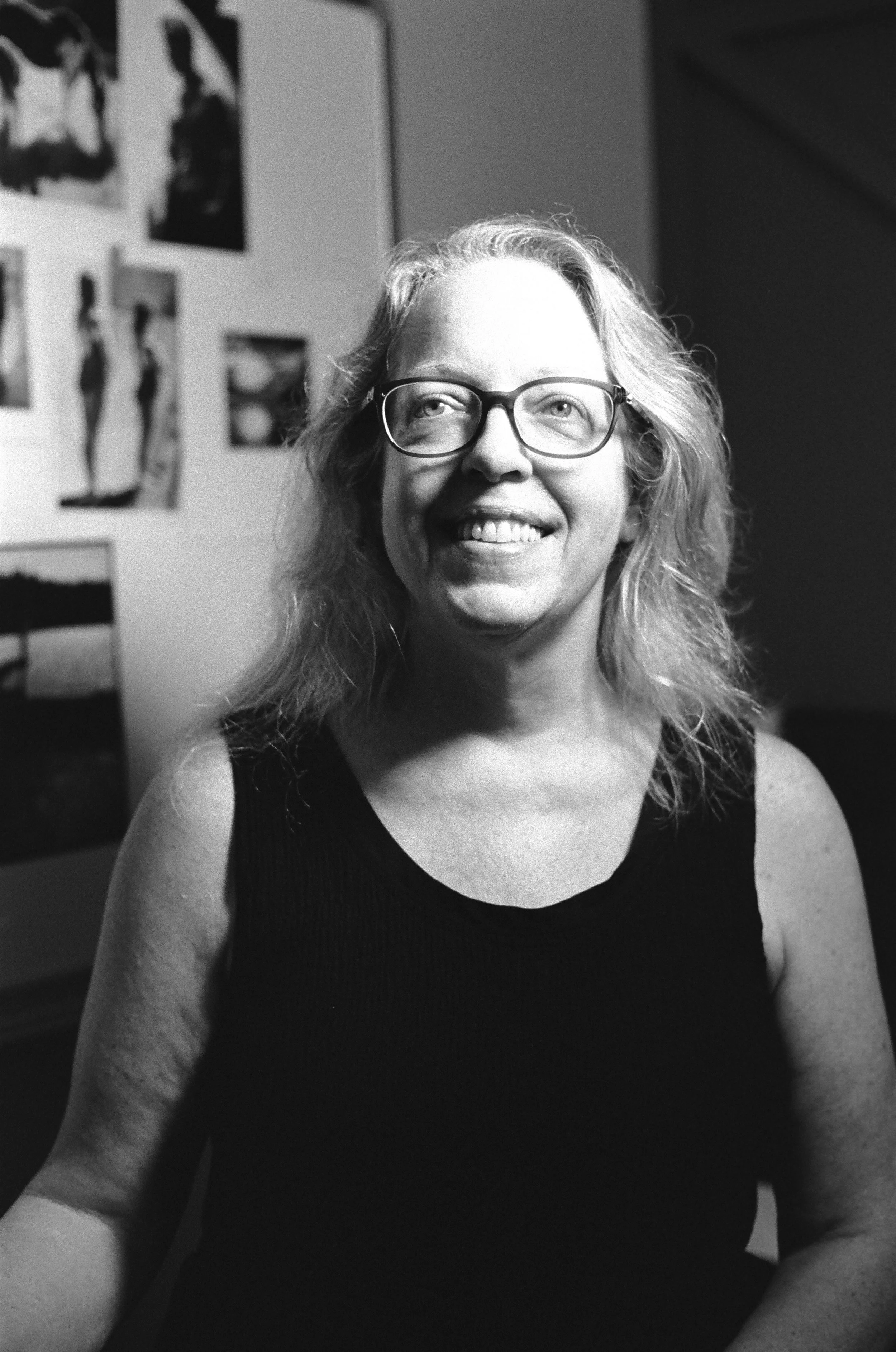 Black and white photo of a woman with glasses smiling, wearing a sleeveless top, sitting in front of a wall with various photographs and artwork.