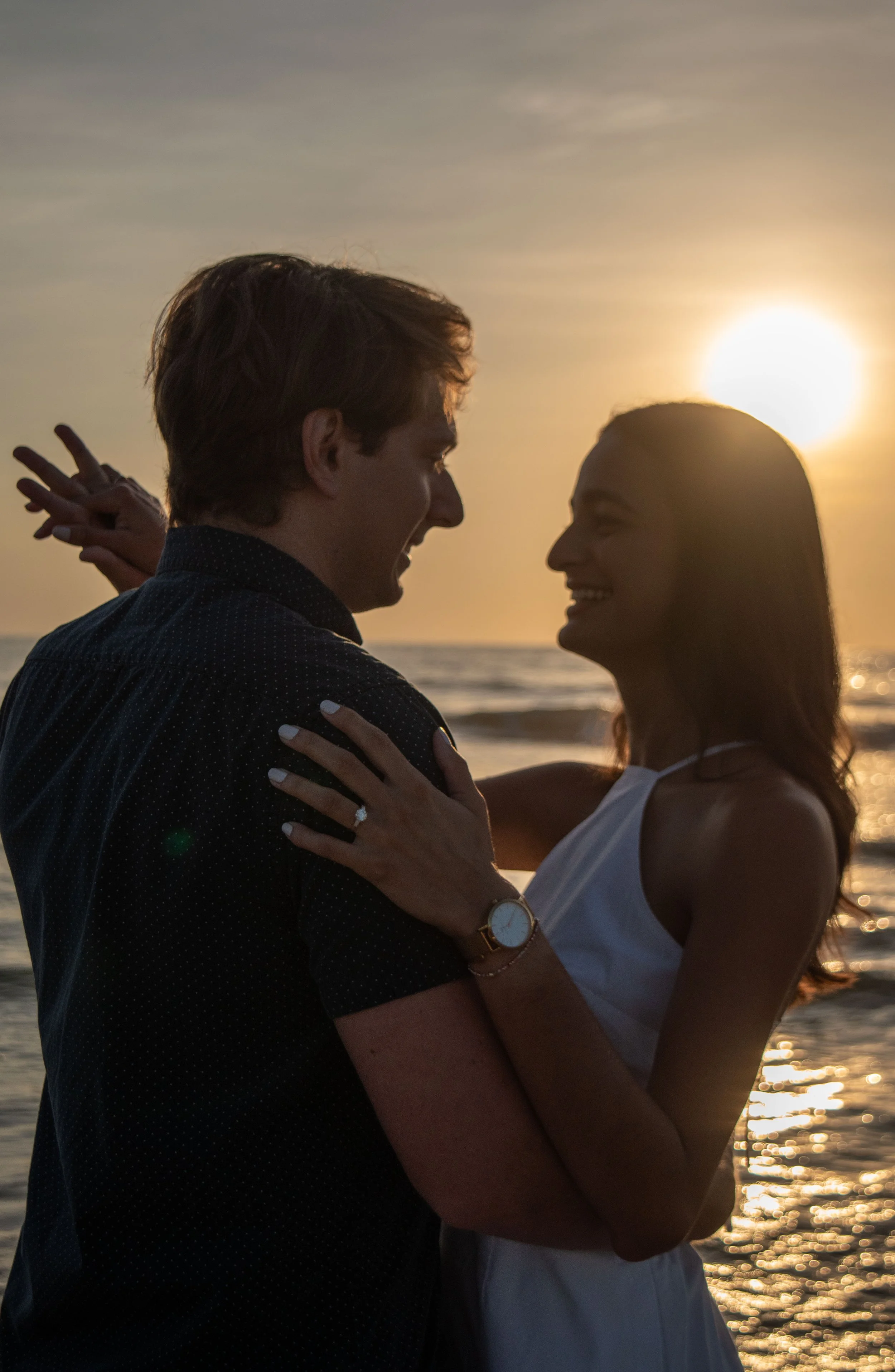 A couple stands close together, smiling and looking into each other's eyes at sunset on a beach, with the sun setting over the water in the background.