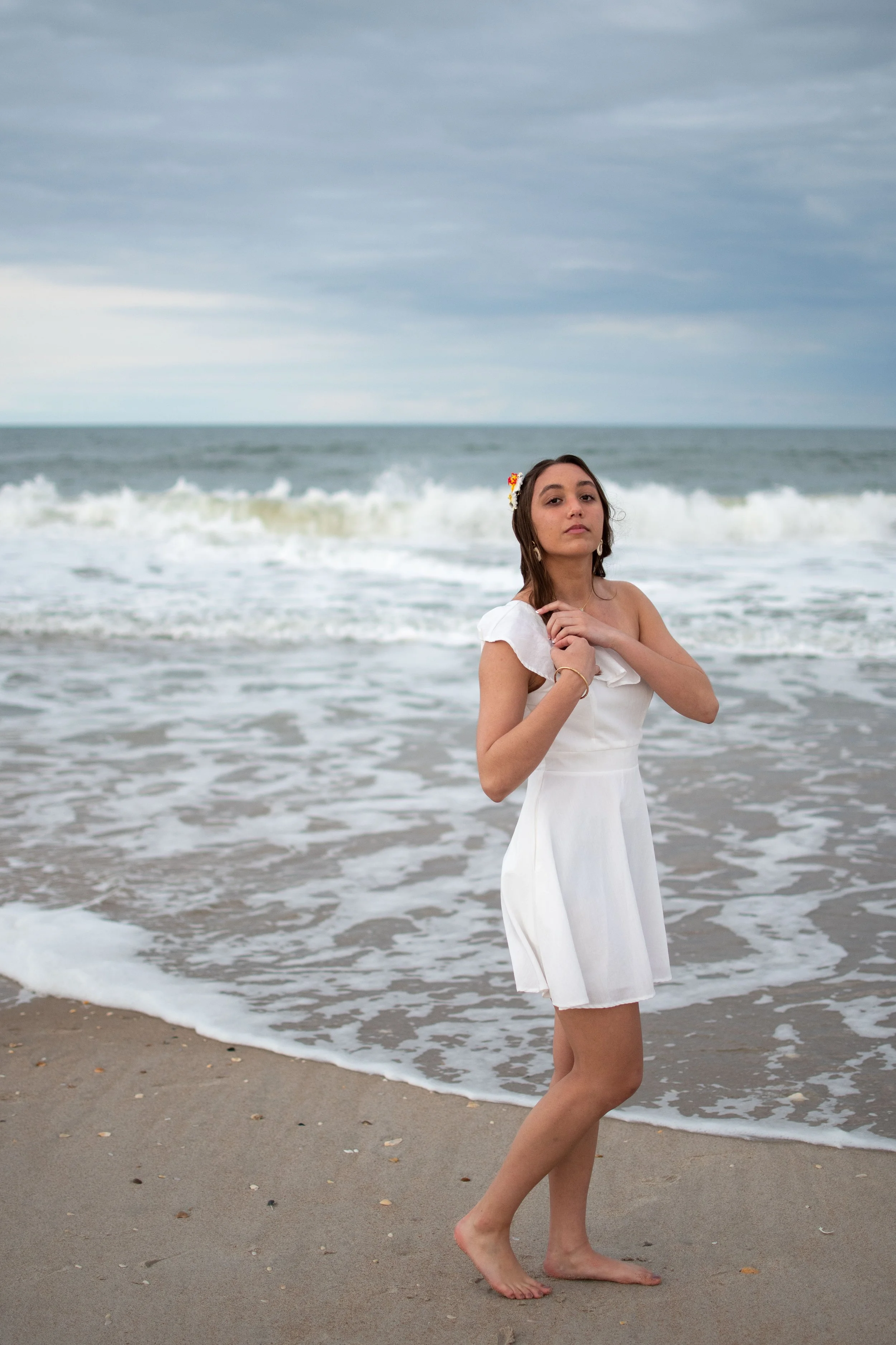 A young woman in a white dress standing barefoot on the sandy beach near the ocean waves, with an overcast sky in the background and wearing floral earrings and a flower in her hair.