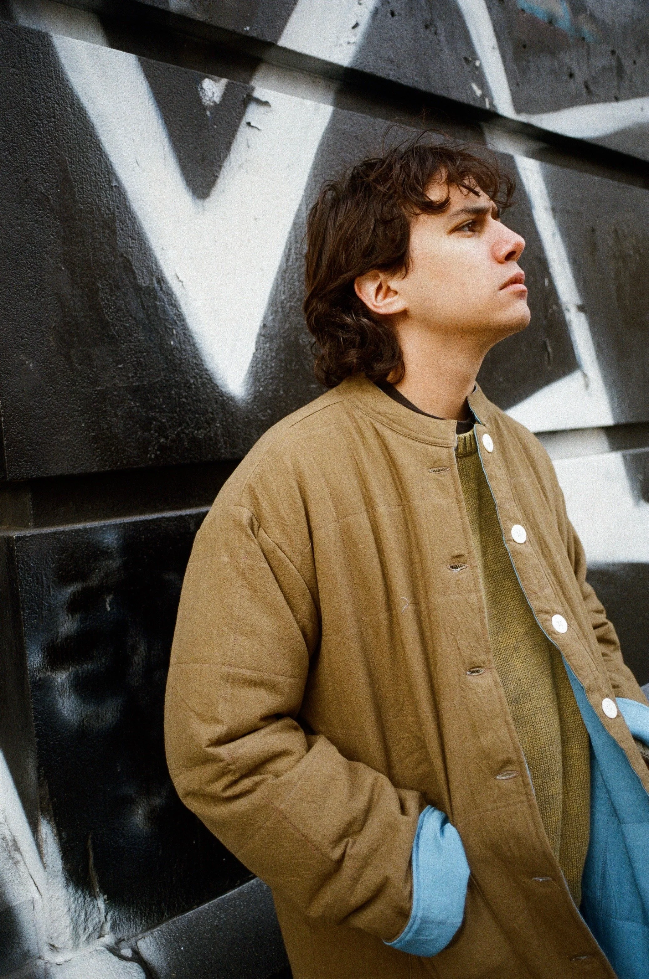 A young man with dark curly hair standing against a graffiti wall with black, white, and gray colors. He is wearing a tan jacket over a green sweater and blue gloves, looking off to the side with a serious expression.