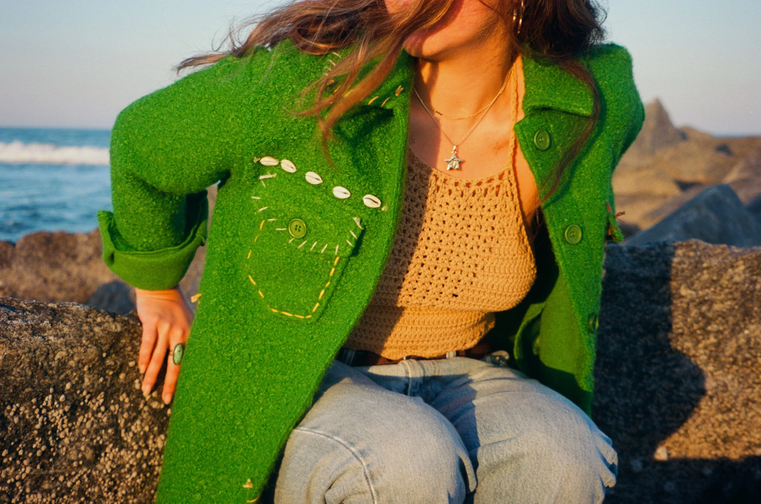 A woman wearing a bright green coat with shell embellishments sitting on rocks near the ocean during sunset.