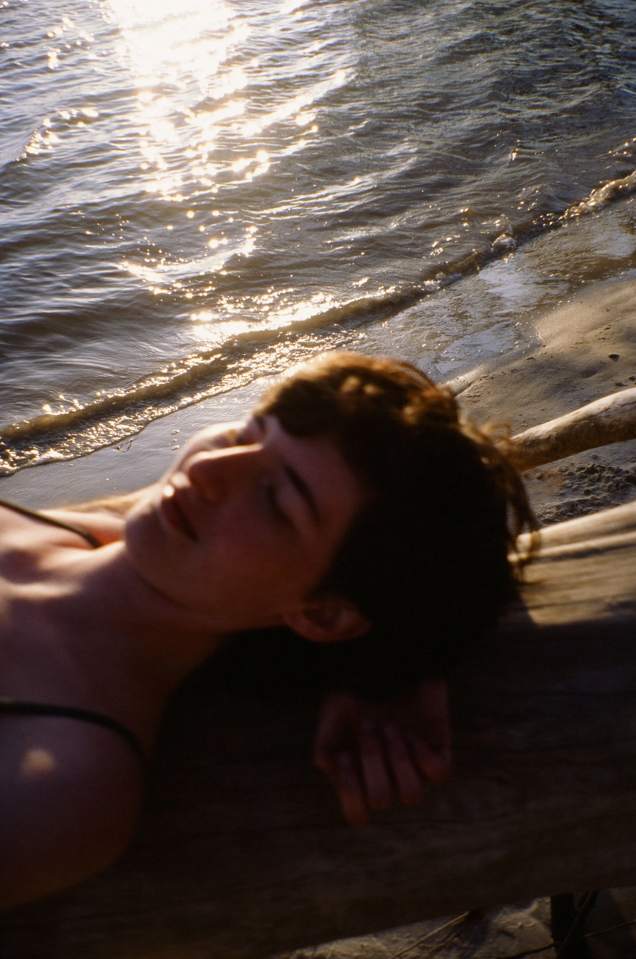 Person lying on a wooden dock near the water with waves gently lapping nearby, during sunset or sunrise.