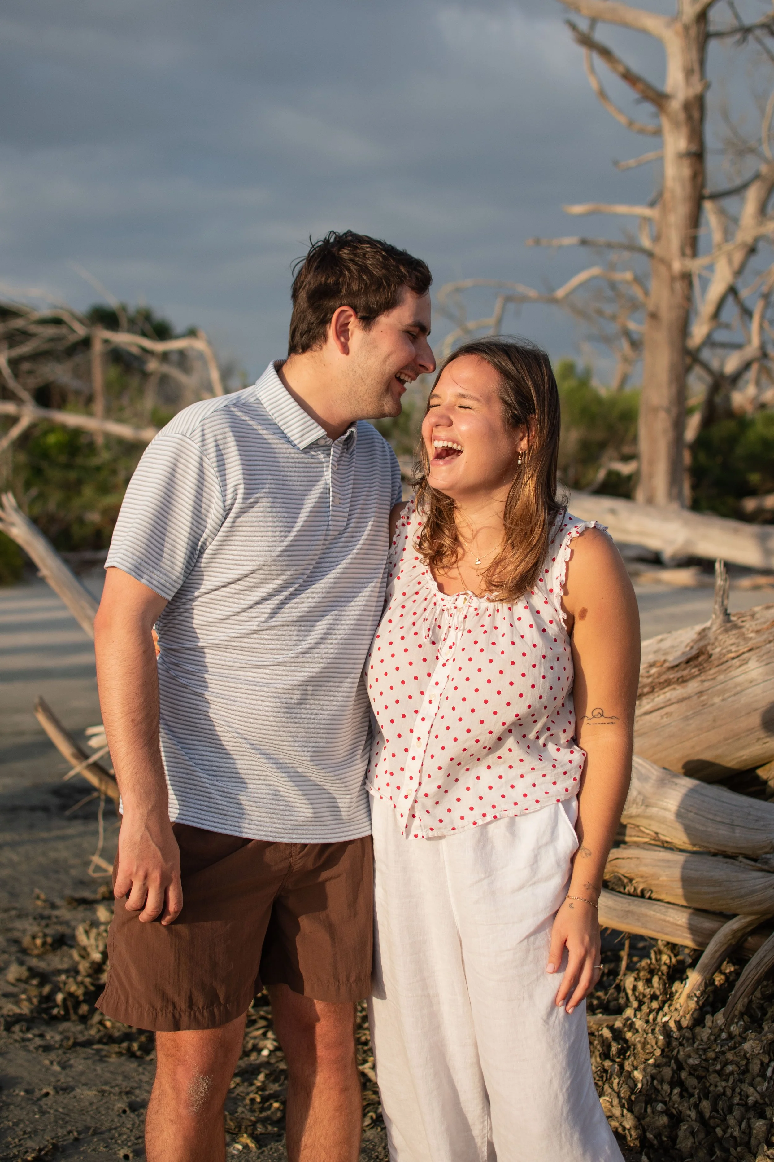 A young couple is laughing and smiling at each other on a beach with driftwood and a cloudy sky in the background.