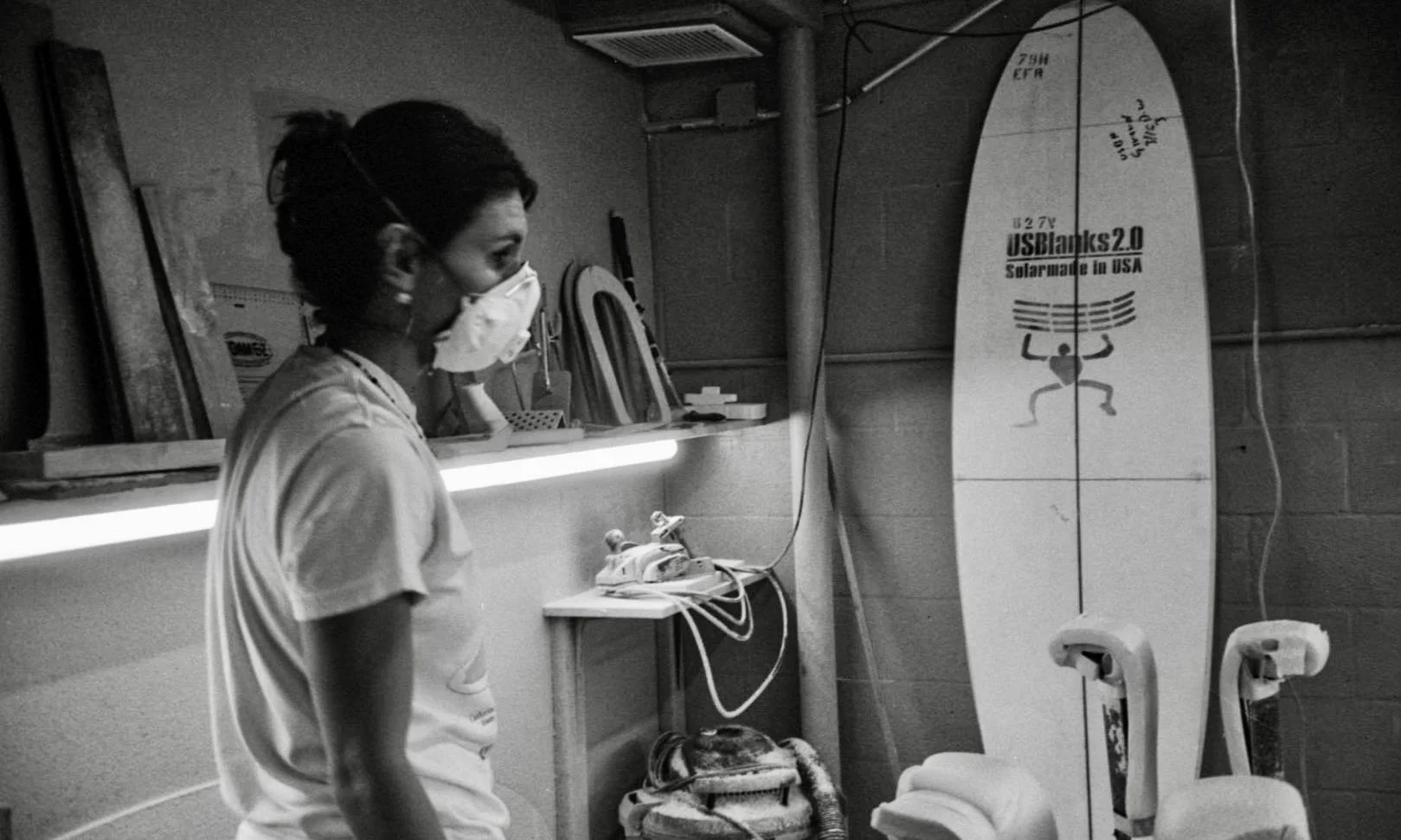 A woman wearing a face mask standing in a room with a surfboard, plastic chairs, and a small shelf with various objects.