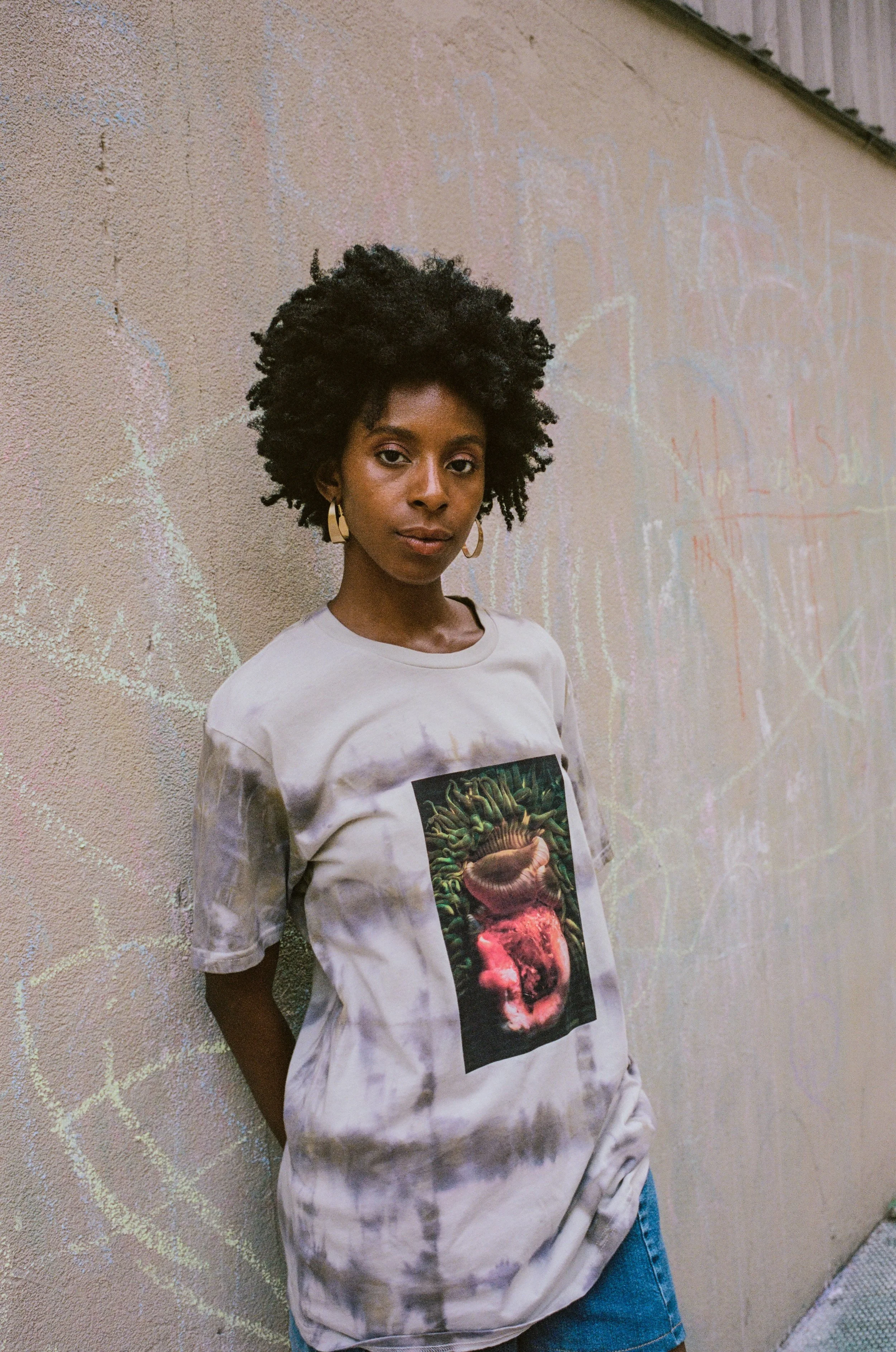 A young woman with dark skin and natural curly hair standing against a textured beige wall with graffiti. She is wearing a light-colored oversized t-shirt with a graphic of a creature and a brain, and denim shorts. She has hoop earrings and a calm ex