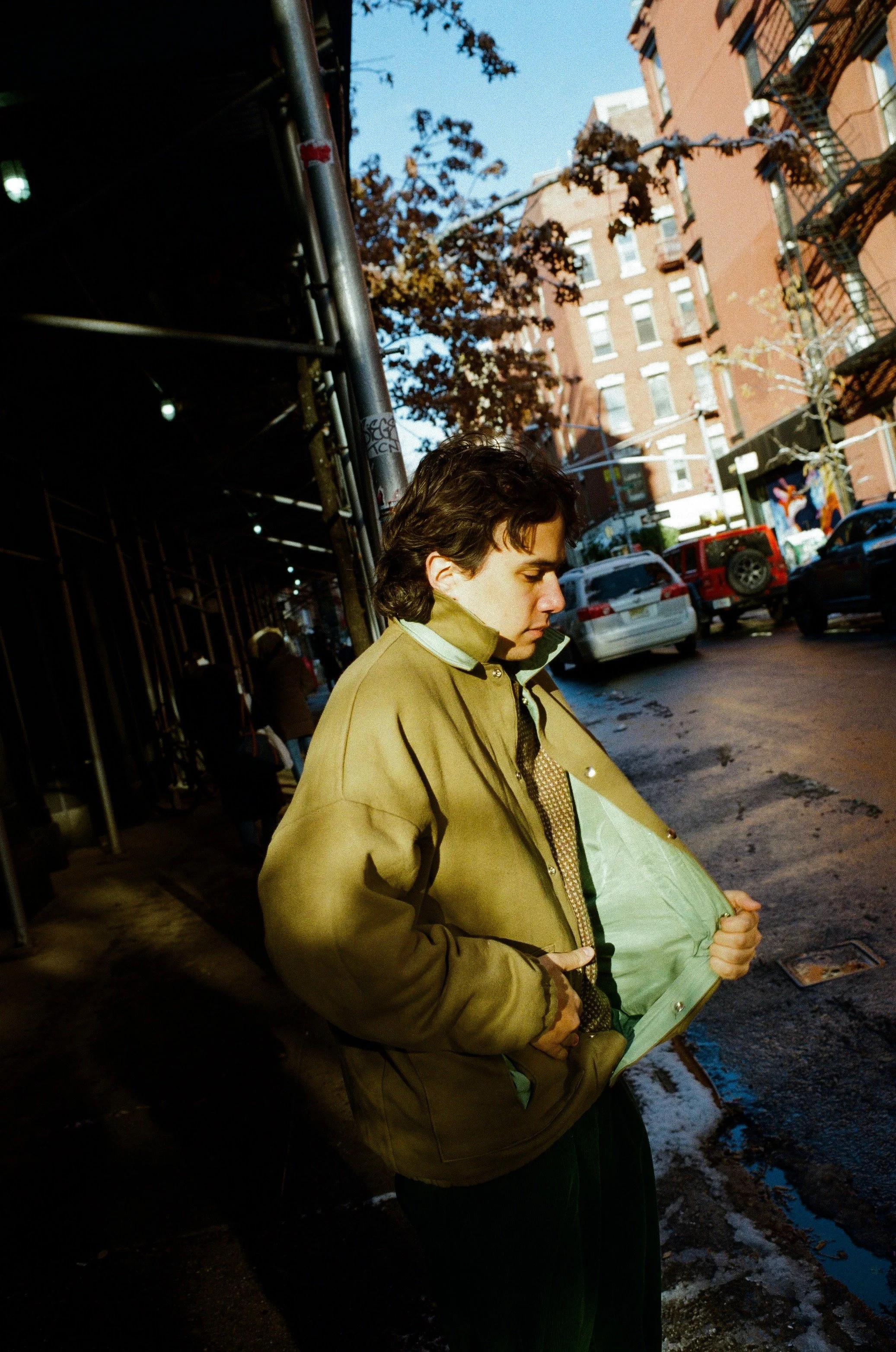A person with dark, wavy hair, wearing a beige jacket, is standing on a city sidewalk with parked cars and a red brick building in the background. The person appears to be looking downward.