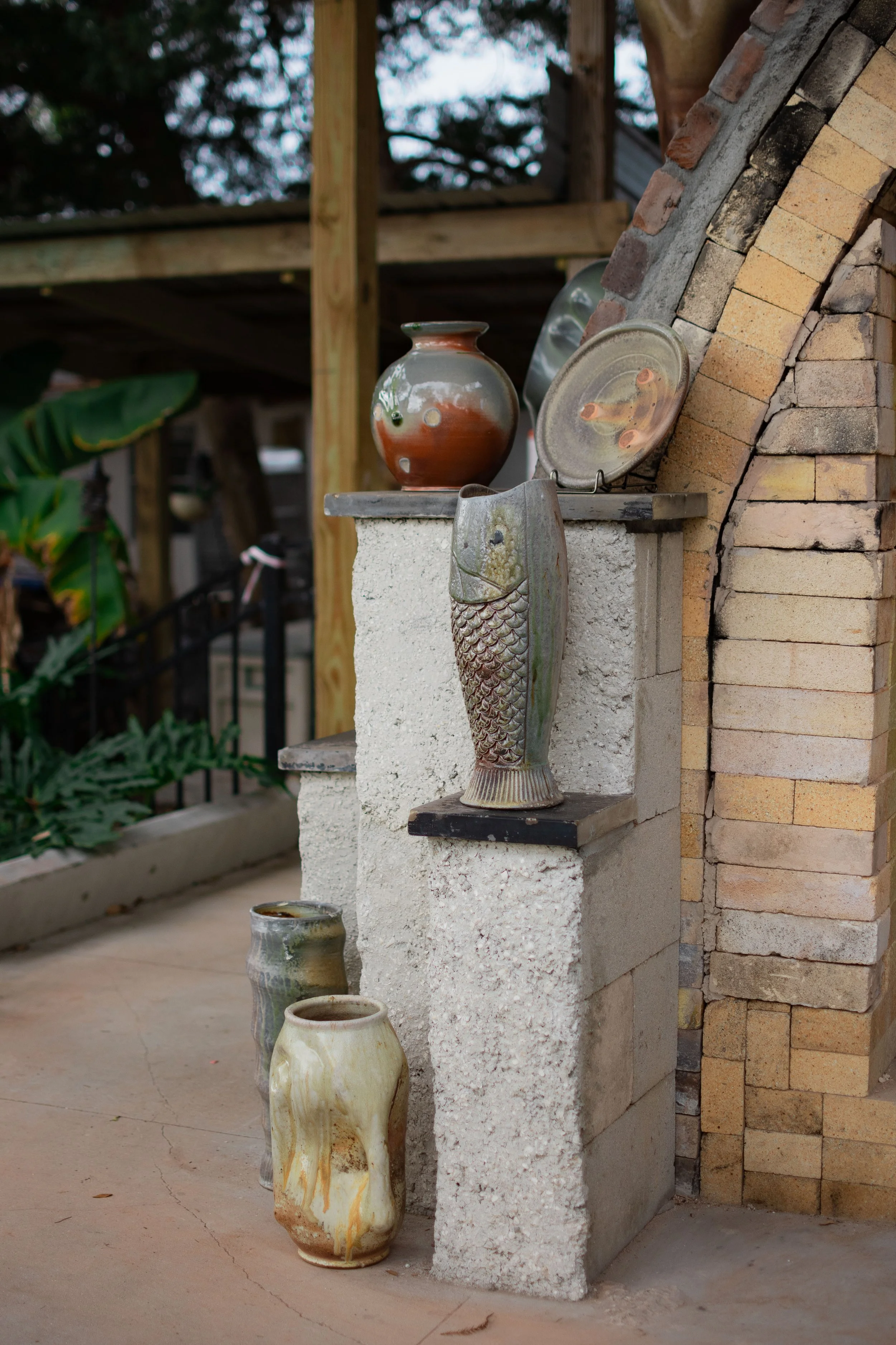 Collection of ceramic vases and platters displayed on a concrete and brick shelf outside, with greenery in the background.