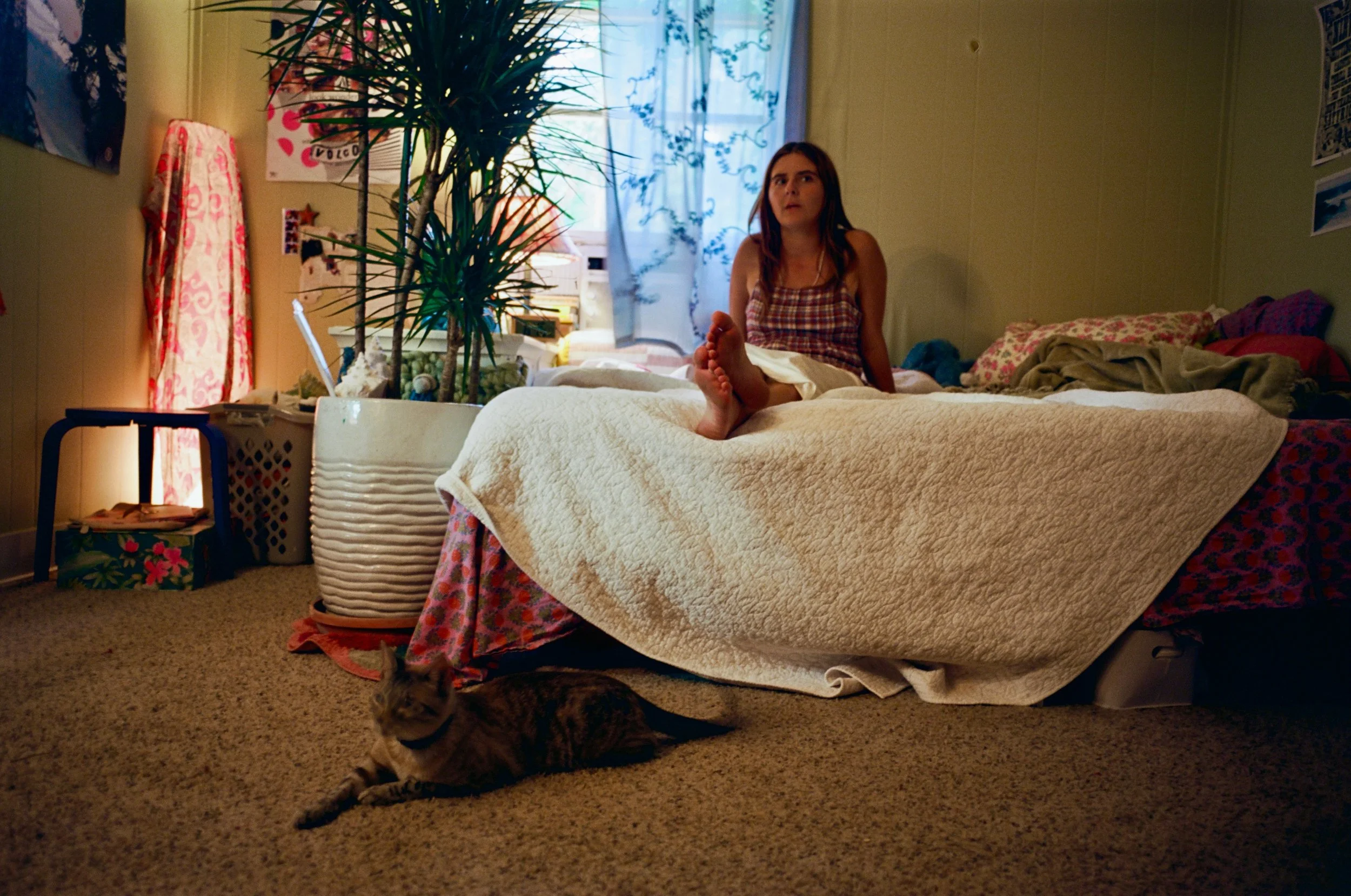 A woman sitting on a bed in a bedroom, looking upward. The bed has a textured blanket and a pink floral skirt hanging from the side. A cat with a striped coat is lying on the carpeted floor near the bed's foot. The room has a large plant in a white p