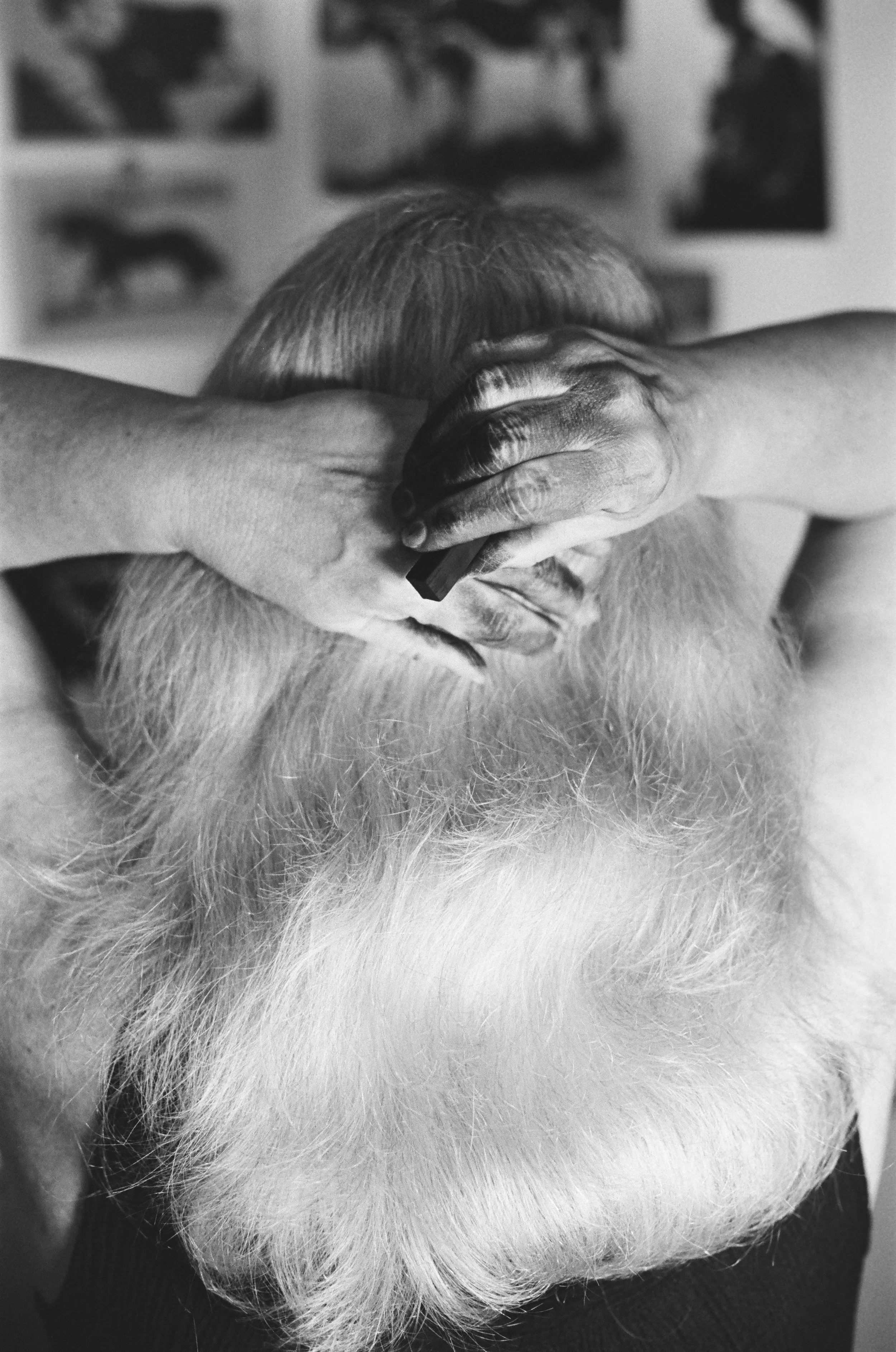 Close-up black and white photo of a person with white hair and beard, resting their head on their arm, with a blurred background of pictures on the wall.