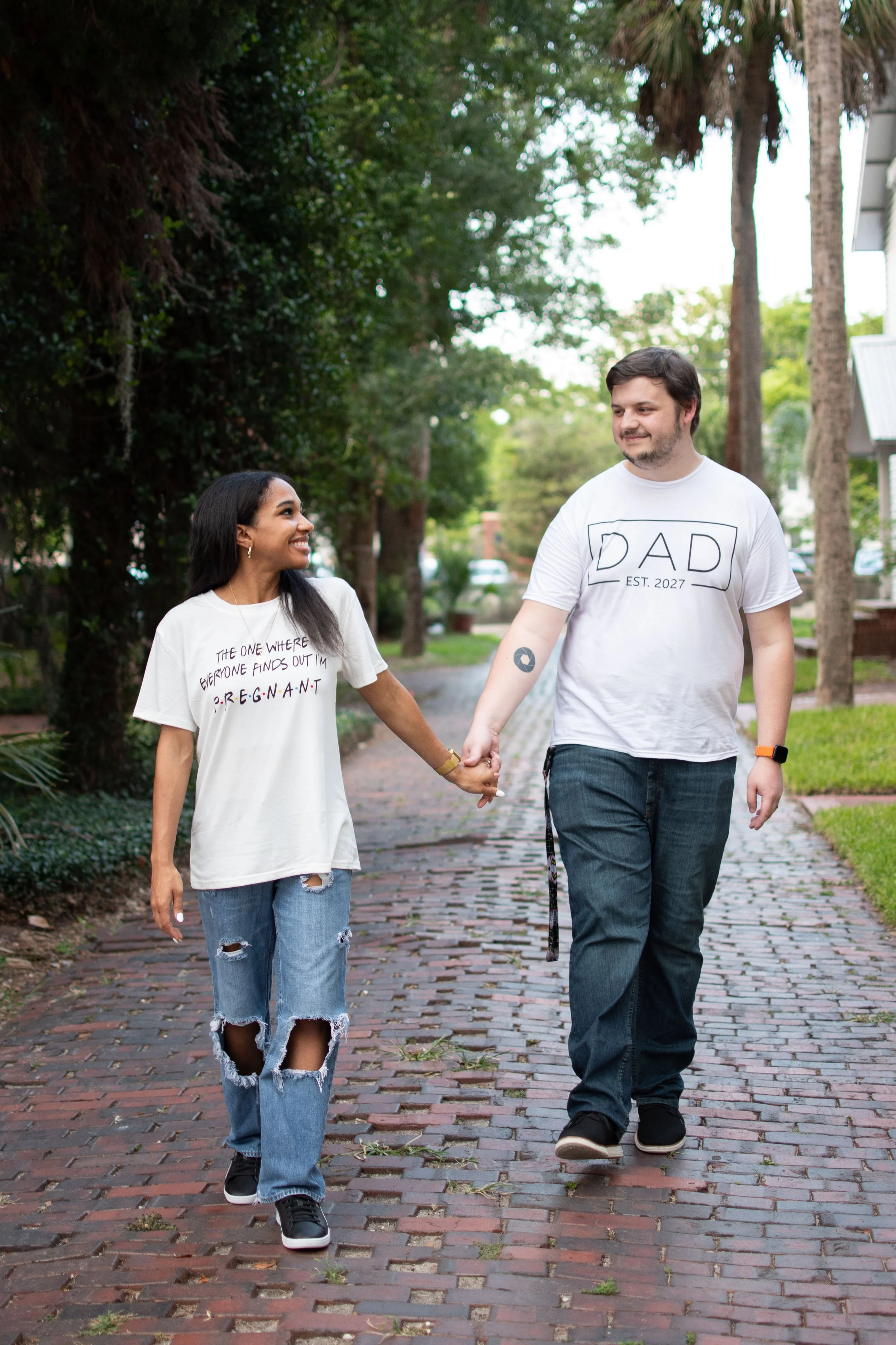 A man and woman holding hands and walking along a brick path in a park. The woman has long black hair, is wearing a white T-shirt with text about pregnancy, ripped jeans, and black sneakers. The man has short dark hair, is wearing a white T-shirt wit