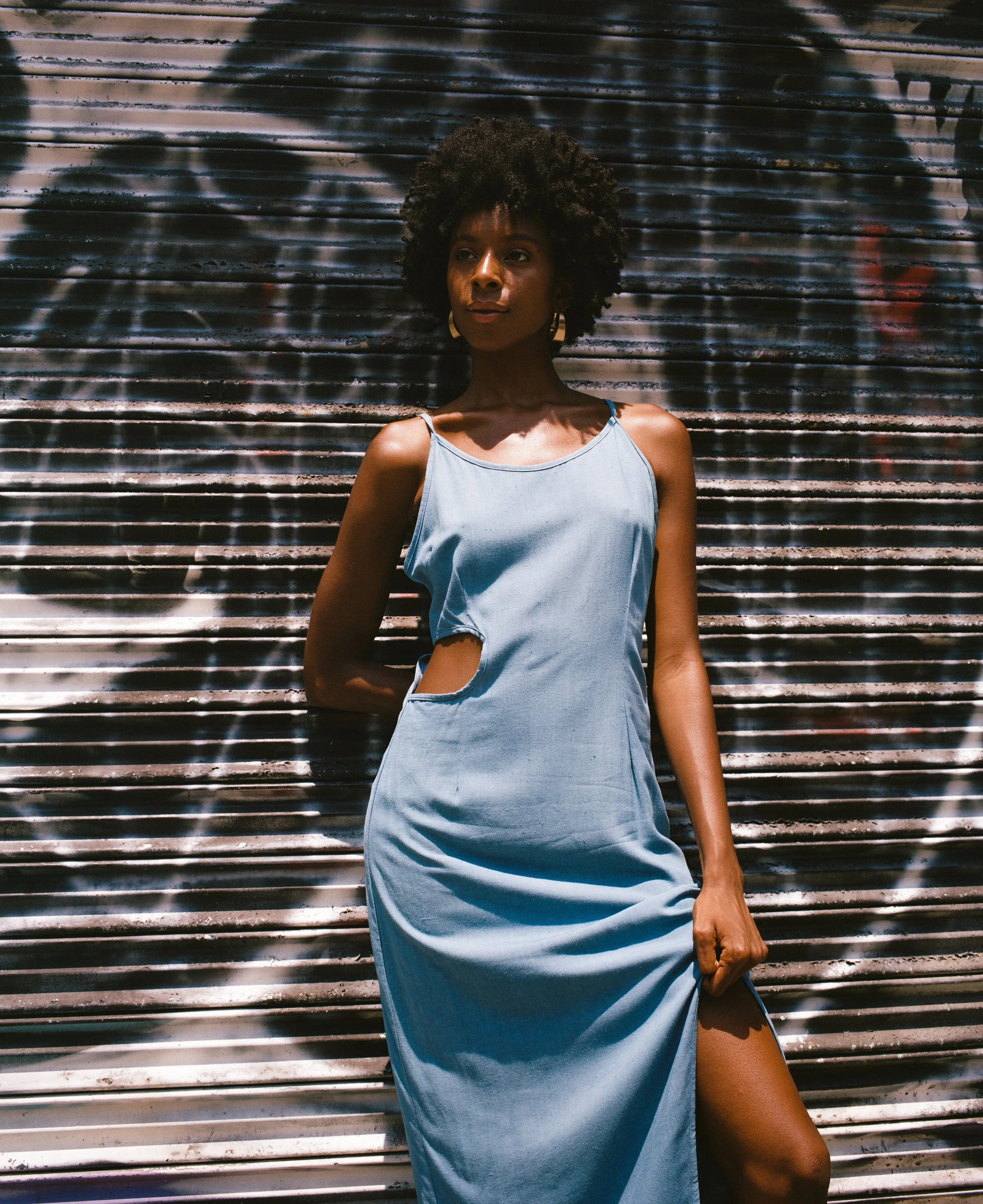 A woman with dark skin and curly hair standing in front of a graffiti-covered metal shutter. She is wearing a light blue sleeveless dress with a cutout on the side and large hoop earrings, looking off to the side with a neutral expression.