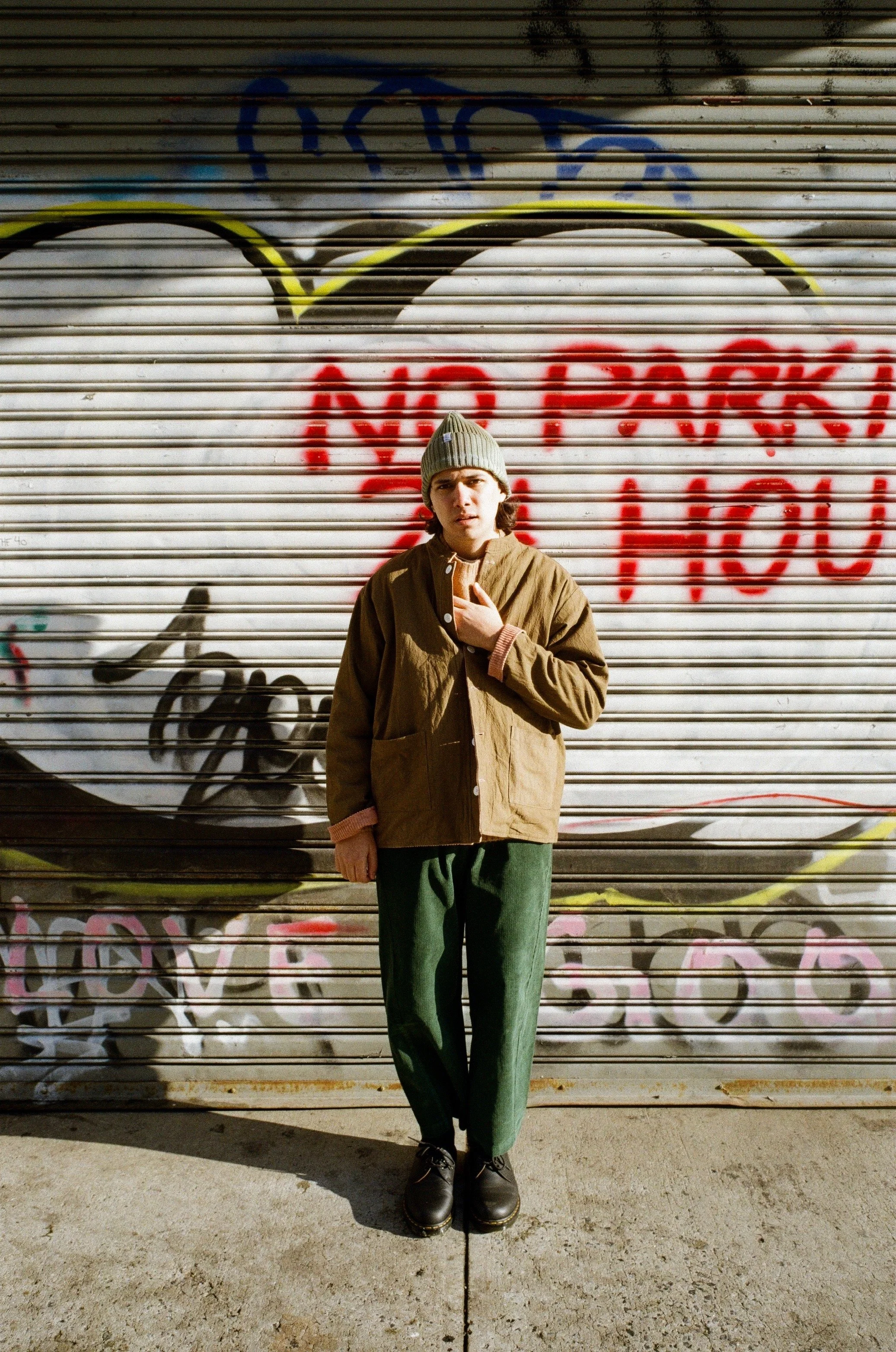 A young person wearing a gray knit hat, brown jacket, and green pants, standing in front of a graffiti-covered metal roll-up door, with one hand on their chest and a serious expression.