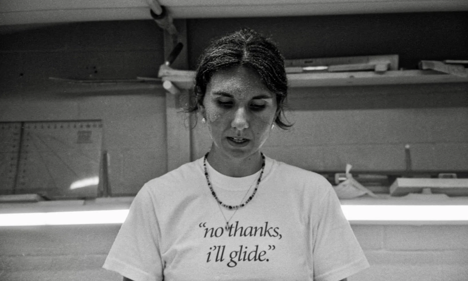 A woman with short hair wearing a white T-shirt that says "no thanks, I'll glide" and a beaded necklace, standing inside a room with shelves, looking down.