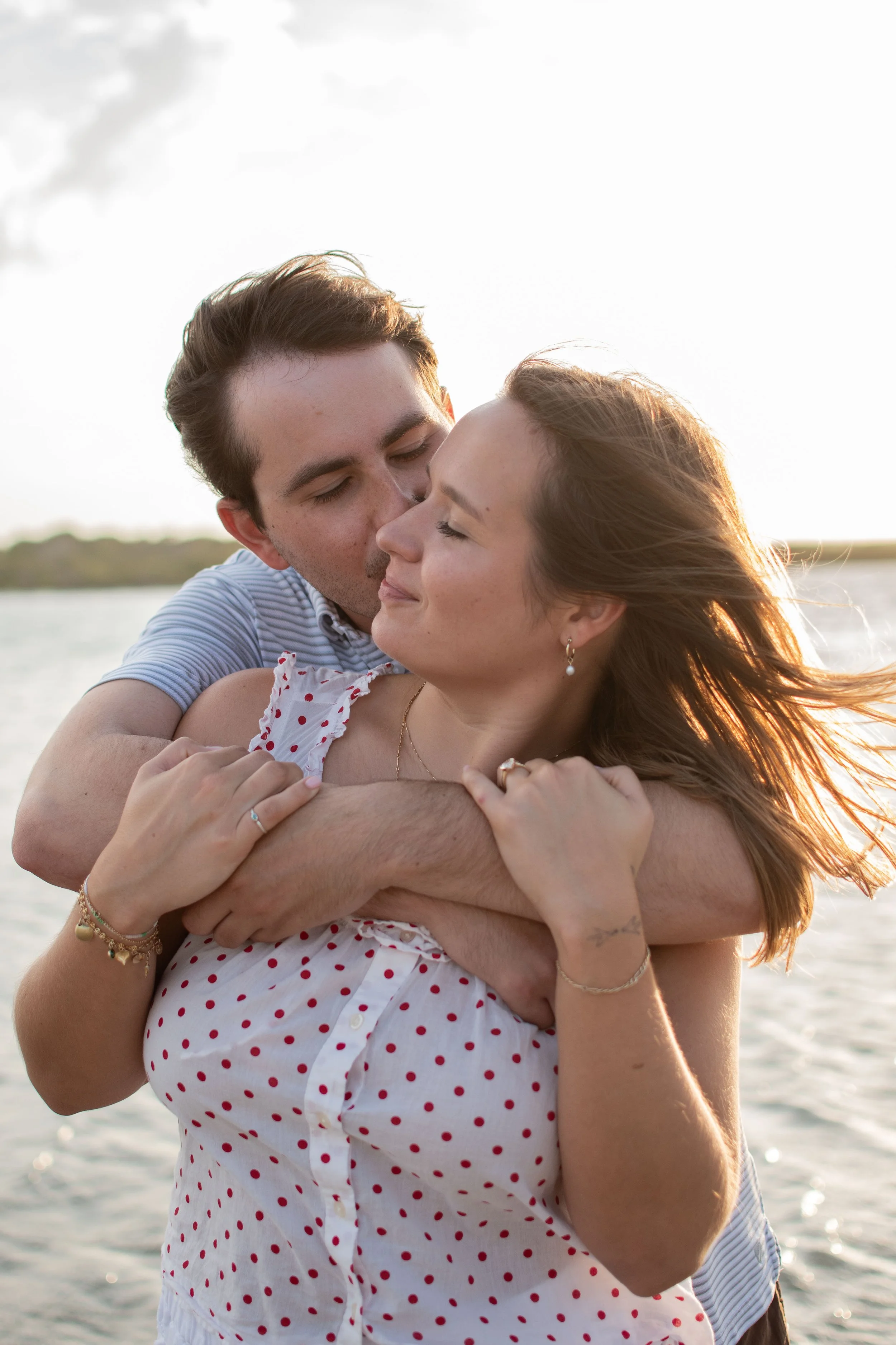 A couple is embracing outdoors near a body of water, with the woman smiling and the man kissing her cheek, during sunset.
