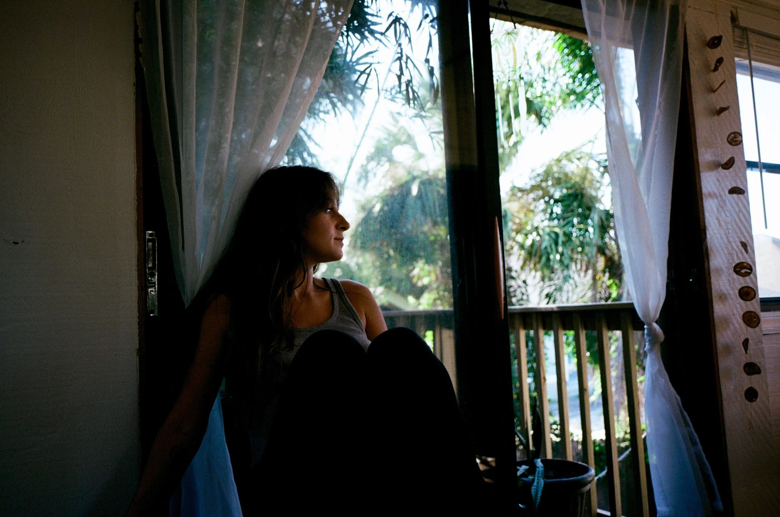 A woman sitting by a window with her profile visible, looking outside at greenery with trees and leaves, near white curtains with decorative round accents on the right side.